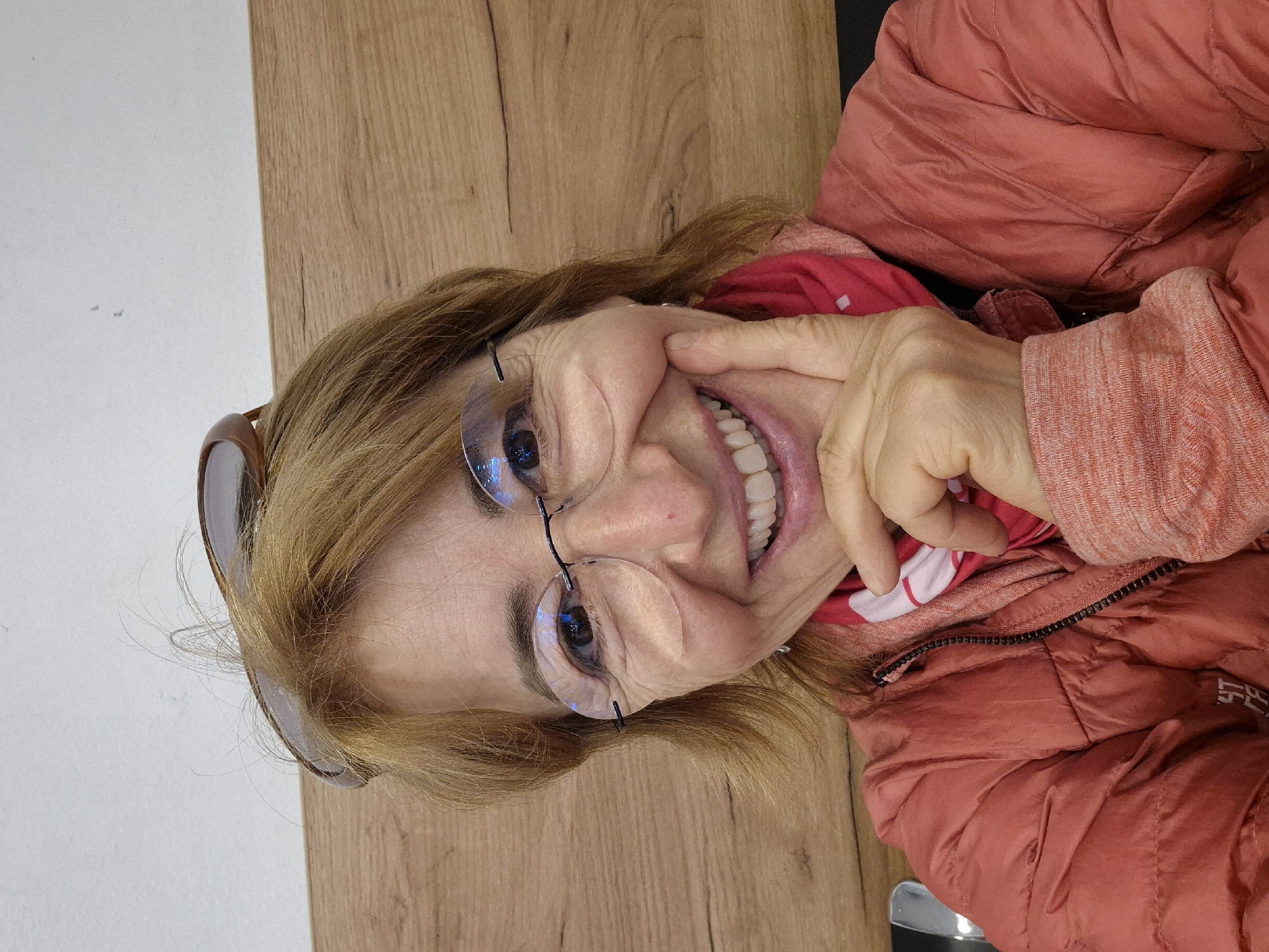 A woman with glasses is smiling and looking at the camera. She is wearing a reddish-orange puffer jacket and a red and white patterned scarf or shirt underneath. Her hair is blonde, and sunglasses are perched on top of her head. She is touching her face with her right hand. The background includes a light-colored wooden surface and a glimpse of a white wall. There is no indication of the location being in Vienna, Austria from the visual information provided.