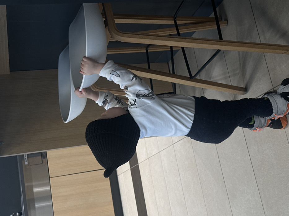 An indoor scene in Ivashevo, Russia, captures a young child standing and facing right, with both hands grasping the front edge of a white, saddle-shaped bar stool seat for support. The child wears a black knitted hat with a pointed top, a long-sleeved white shirt patterned with dark shapes, black textured pants, and grey and orange winter boots. To the child's left and in the background, a light wood counter with a darker surface is visible, above which a stainless steel appliance, possibly a water cooler or coffee machine, is partially seen. The floor is composed of light-colored rectangular tiles. Two identical bar stools, featuring light wooden frames and black metal footrests, are positioned to the right. The immediate background behind the stools consists of a dark, uniform vertical panel or wall. The environment appears to be a modern commercial or public space, such as a cafe or shared lounge area.