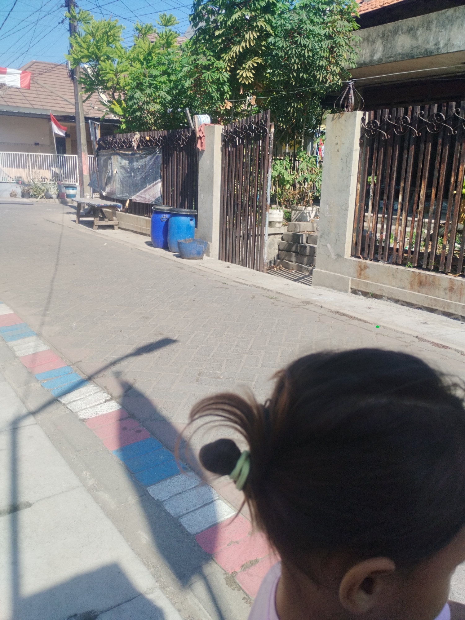 The image depicts a street scene in the city of Surabaya, Indonesia, on a clear, sunny day. In the foreground, the back of a person's head is visible, with dark hair tied in a ponytail with a green elastic band. The person is wearing a light-colored top. The street is paved with interlocking bricks and has a colorful border along the sidewalk, featuring red, white, and blue painted rectangles. A shadow, possibly from a lamppost or overhead structure, stretches across the pavement.
To the left of the street, a house with a brown tiled roof and a white fence is visible, with an Indonesian flag (red and white) displayed. Adjacent to the house, a wooden table and two blue plastic barrels are placed near a metal gate and a wall. Behind the gate, trees with green foliage are prominent. A concrete pillar topped with an ornamental metal structure stands near the gate. The overall environment appears to be a residential area with some utility poles and overhead wires visible in the background.