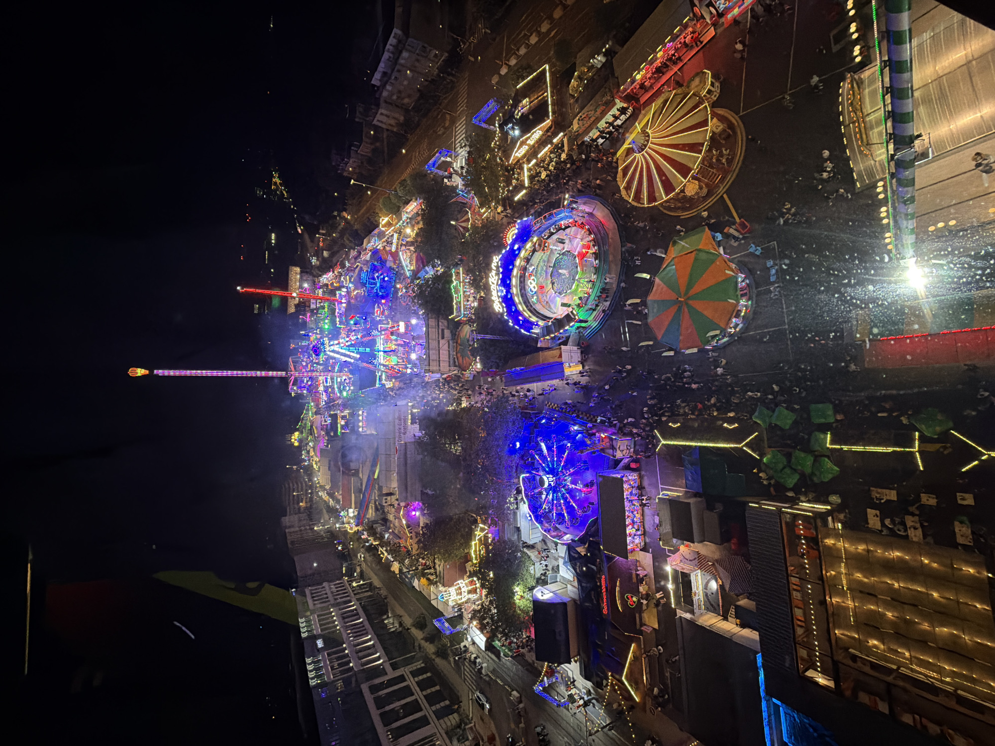 This is an aerial, nighttime view of a fairground located in the city of Luxembourg, Luxembourg. The fairground is filled with numerous amusement rides, illuminated by a vibrant array of colorful lights. In the background, several multi-story buildings are visible, suggesting an urban setting. The foreground shows a section of the fairground with various attractions, including a carousel with red and yellow striped awnings, a circular ride with blue lighting, and a tent-like structure. Many people are present, dispersed throughout the fairground, walking between rides and congregating in various areas. The overall scene depicts a lively and bustling atmosphere typical of a public fair.