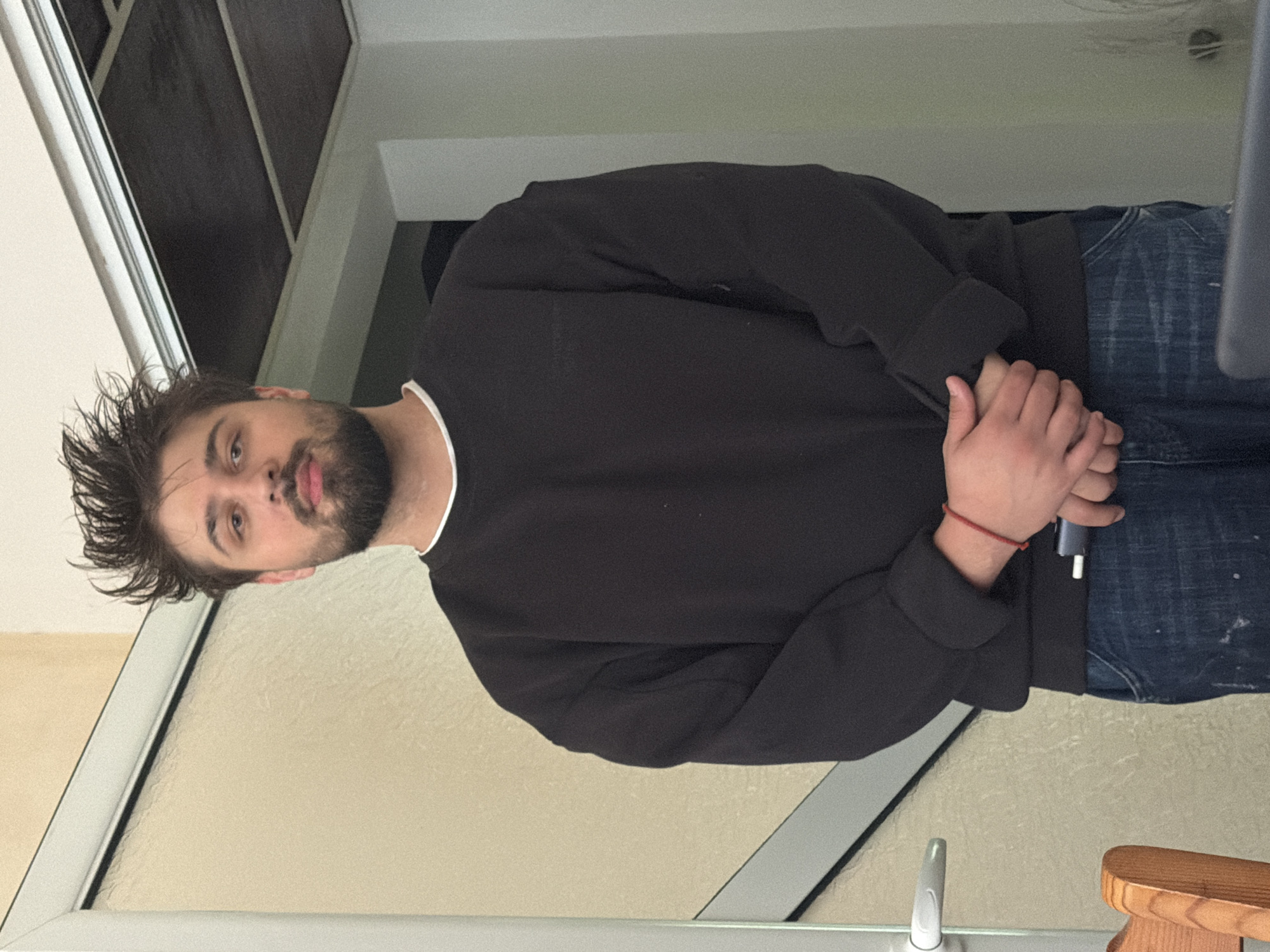 A man with short, dark hair and a beard is standing indoors, wearing a black sweatshirt and blue jeans. He is holding something in his hands, possibly a small device, and his fingers are interlocked. He is standing next to a glass door or window frame, with a white trim. A portion of a wooden stool or chair is visible in the lower right corner. The location is indoors, with a blurred background. The location in the city of Varna, Bulgaria cannot be determined from the given information.