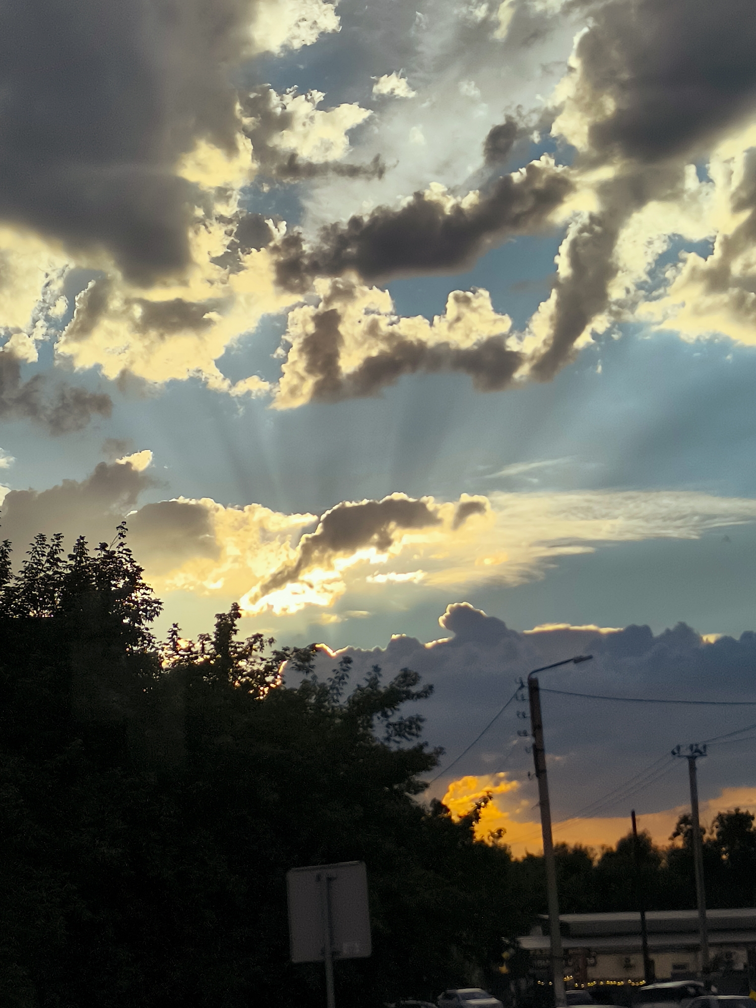 A low-angle, vertical shot captures a dramatic sky over a semi-urban landscape. The sky is filled with a mix of dark, heavy clouds and brighter, illuminated clouds, with shafts of sunlight piercing through. The upper portion of the image is dominated by these clouds. Towards the bottom, silhouetted trees line the left side, partially obscuring a street scene. On the right side of the frame, two utility poles with wires are visible, along with the suggestion of buildings and vehicles in the background. The overall impression is of a dynamic, possibly stormy or sunset-lit sky. The location is Qarağandı city administration, Kazakhstan.