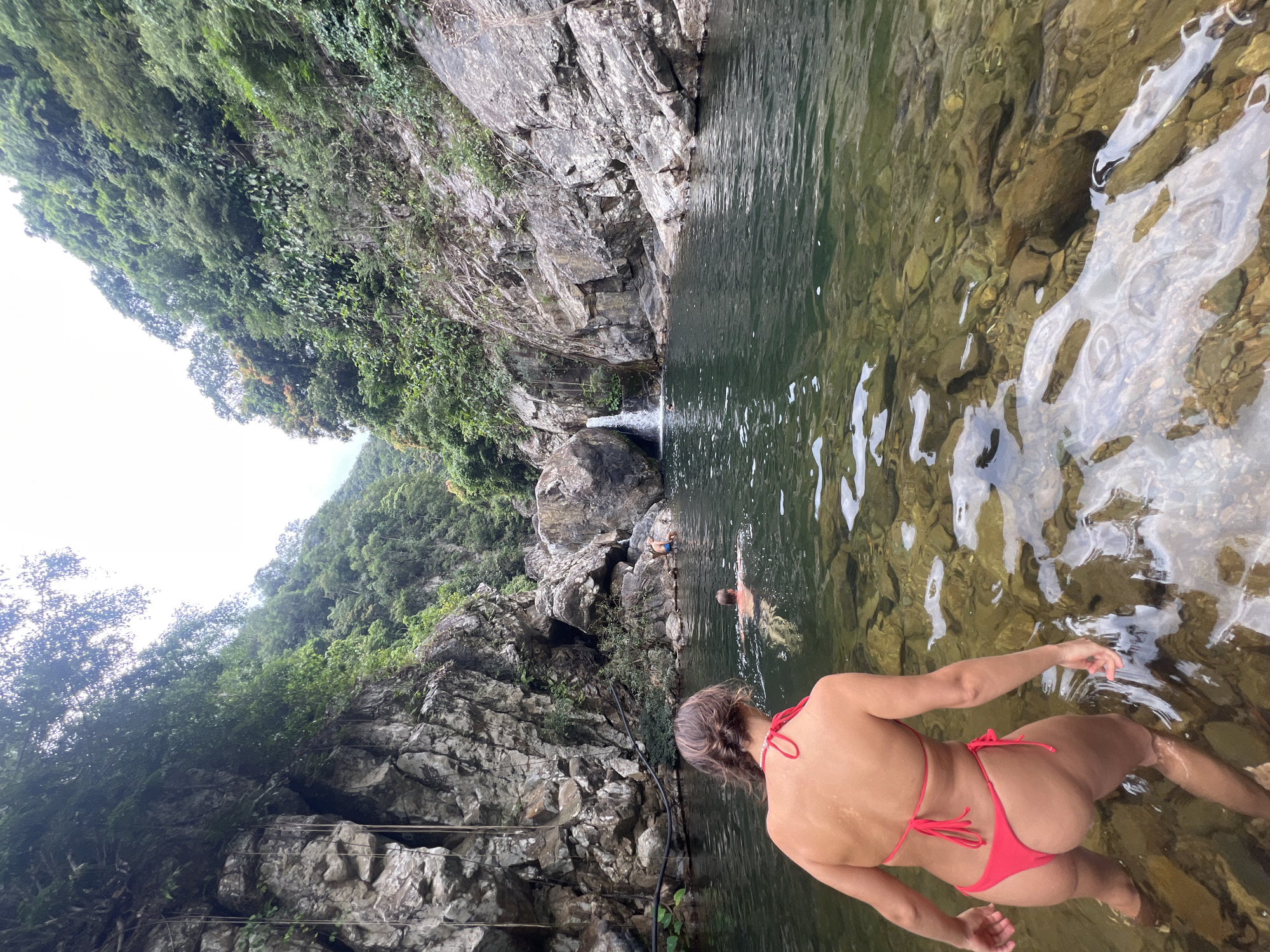 A woman in a pink bikini stands in the shallows of a clear river with a rocky bed. She is facing away from the camera, looking towards the water where two men are swimming. The river is surrounded by lush green trees and rocky cliffs. In the background, a small waterfall cascades into the river. The location is likely a natural swimming spot in the suburban or rural areas of Đà Nẵng, Vietnam, possibly in a district like Hòa Vang or Sơn Trà, known for their natural landscapes and proximity to mountains.