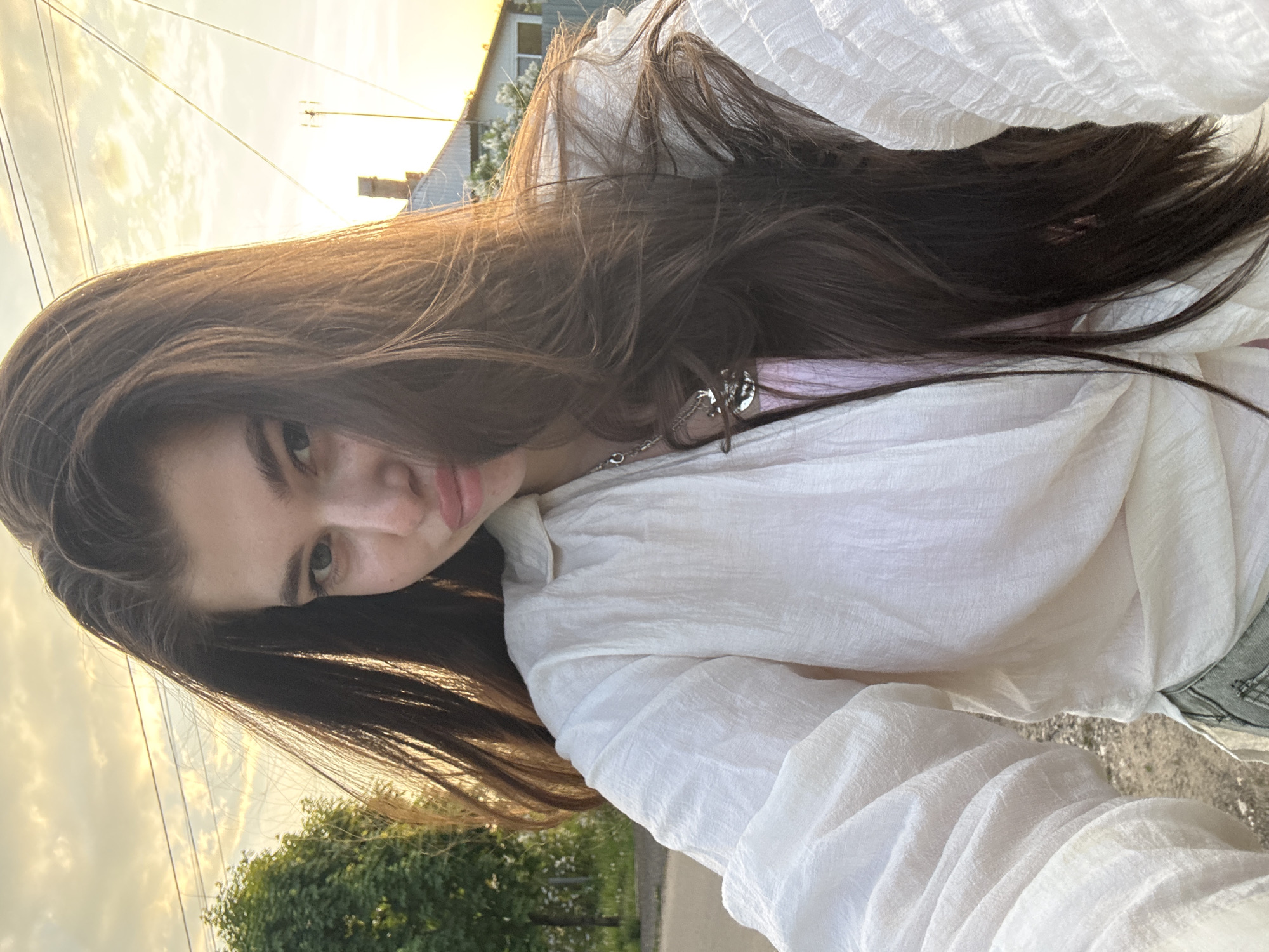 A woman with long brown hair and light skin is seen from a low angle, looking at the camera. She is wearing a white, textured long-sleeved shirt and a necklace. The background shows a partially cloudy sky at sunset with residential buildings and utility lines. Trees and a paved surface are also visible. The location appears to be in a residential area. It is not possible to confirm the location as Мещёры, Russia from the given image.
