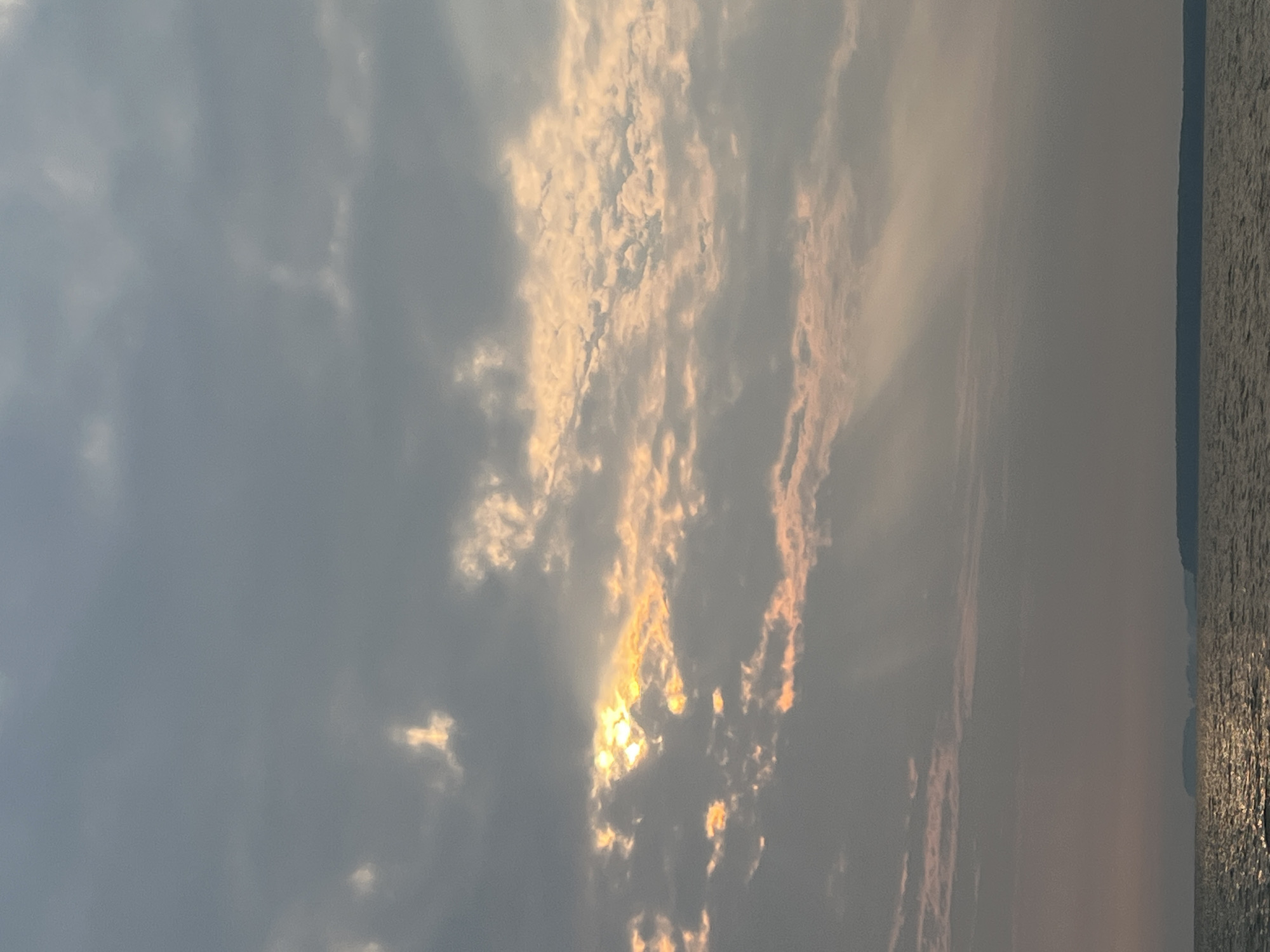 A wide shot captures a serene, overcast sky with the sun peeking through a break in the clouds, casting rays onto the water below. The scene is set on the Sea of Azov, near the shore of the city of Novopichugovo, Russia. The water is calm, reflecting the muted light of the sky, and a distant landmass is visible on the horizon. The overall atmosphere is one of peaceful solitude.