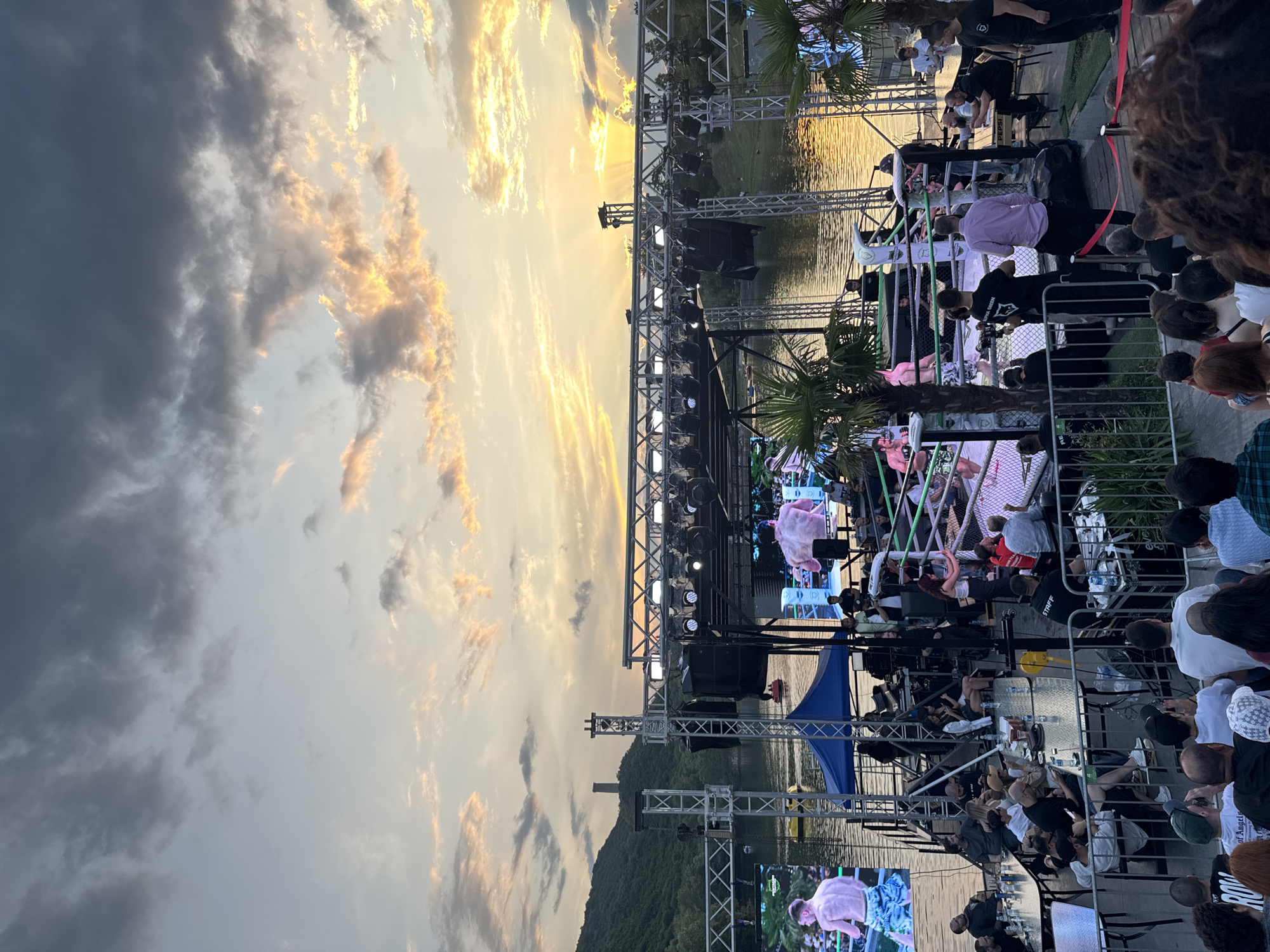 The image depicts an outdoor event, likely a fight or performance, taking place on a platform over a body of water, with a sunset in the background. The platform is equipped with a stage, lighting rigs, and large screens displaying images of athletes. A boxing ring is set up on the platform, with two men in fighting attire visible inside. Spectators are seated at tables on the platform and in an area in the foreground, observing the event. The environment includes palm trees, a hilly landscape with a structure resembling a tower on a hill, and boats on the water. The sky is filled with dramatic clouds illuminated by the setting sun. The scene appears to be in a venue that combines a waterfront location with entertainment facilities. Given the visual cues of the landscape and the unique setting, this event is taking place at the Tbilisi Sea.