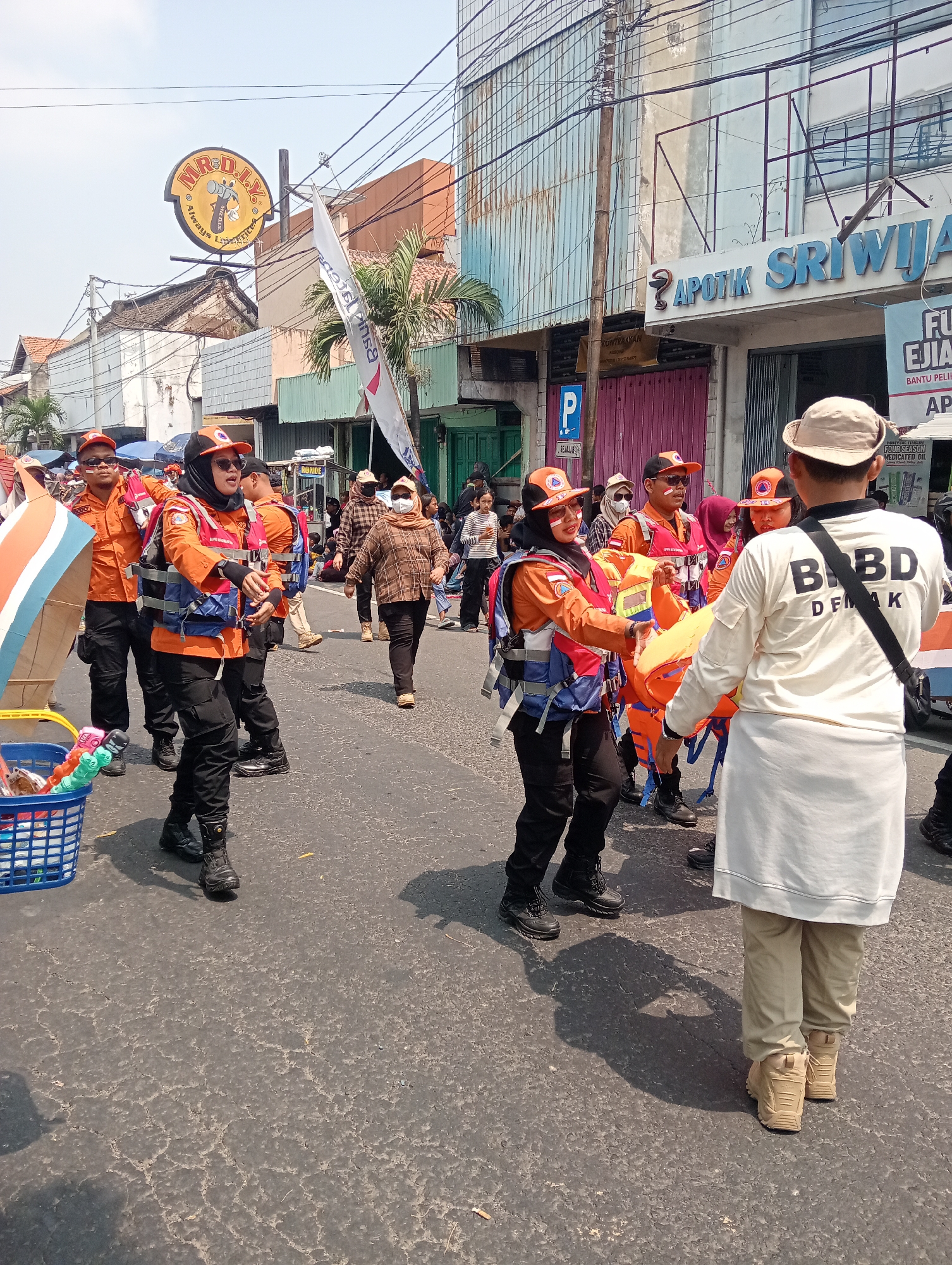 In Demak, Indonesia, a group of individuals in orange uniforms and life vests are participating in a parade or event on a sunlit street lined with buildings. Several people are wearing orange caps and sunglasses. Some are carrying a yellow stretcher with an orange blanket, suggesting a medical or rescue theme. A blue basket filled with items is visible in the foreground. The backdrop includes a store sign that reads "APOTIK SRIWIJA" and another with "MR. D.I.Y". The street surface is asphalt with visible cracks. The overall scene appears to be an outdoor public gathering under a clear sky.