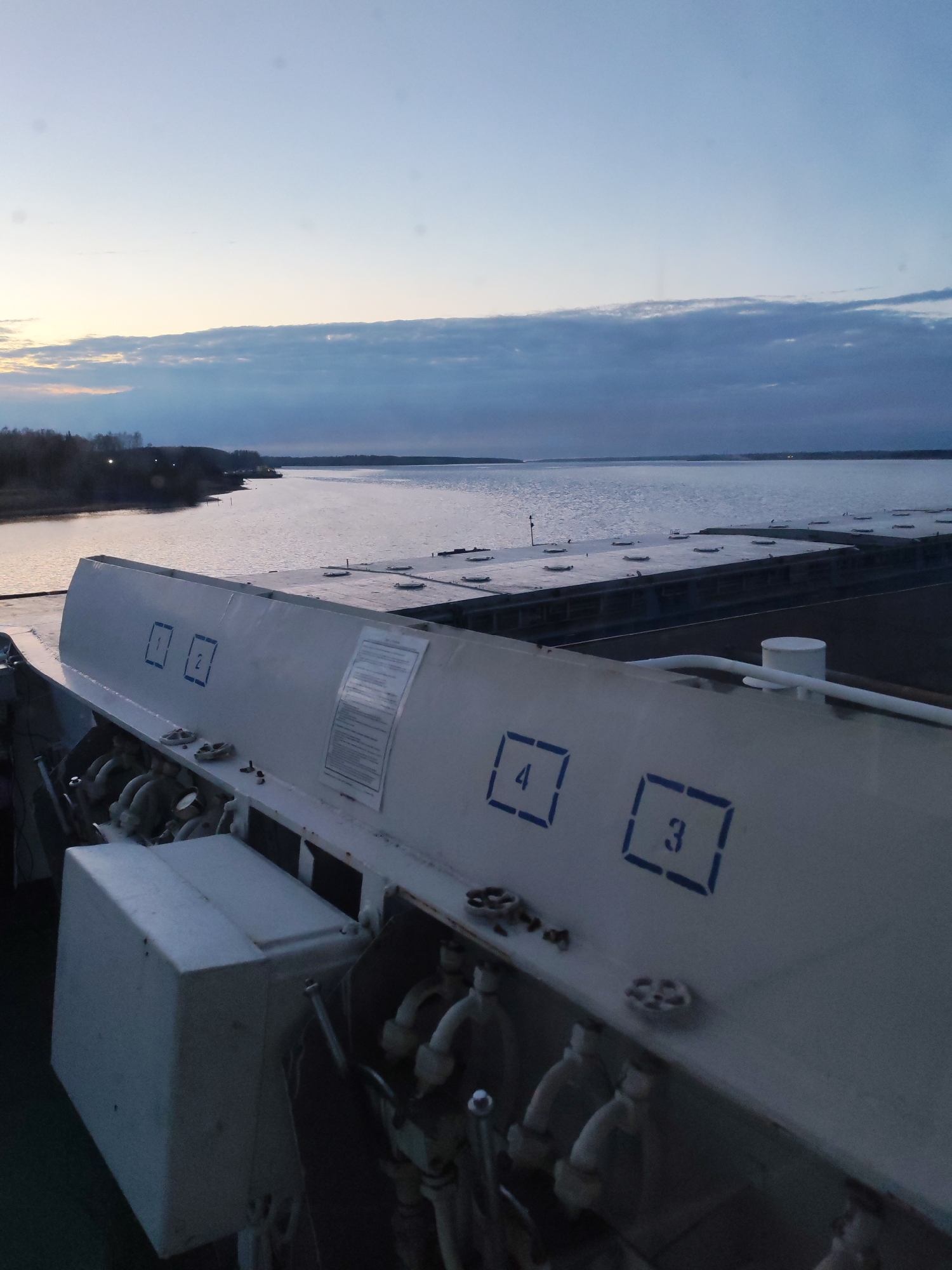 This image shows a view from what appears to be a barge or ship, possibly in the Rybinsk area of Russia, given the body of water and the flat, low-lying land in the distance. The sky suggests either dusk or dawn, with the horizon line containing land with sparse trees.
In the foreground, there is a piece of industrial equipment, most likely part of the barge. It is painted white and features numbered squares in blue with the numbers "1", "2", "4", and "3". There are pipes and valves beneath the numbered panel, indicating some kind of hydraulic or fluid control system. The equipment appears to be made of metal.