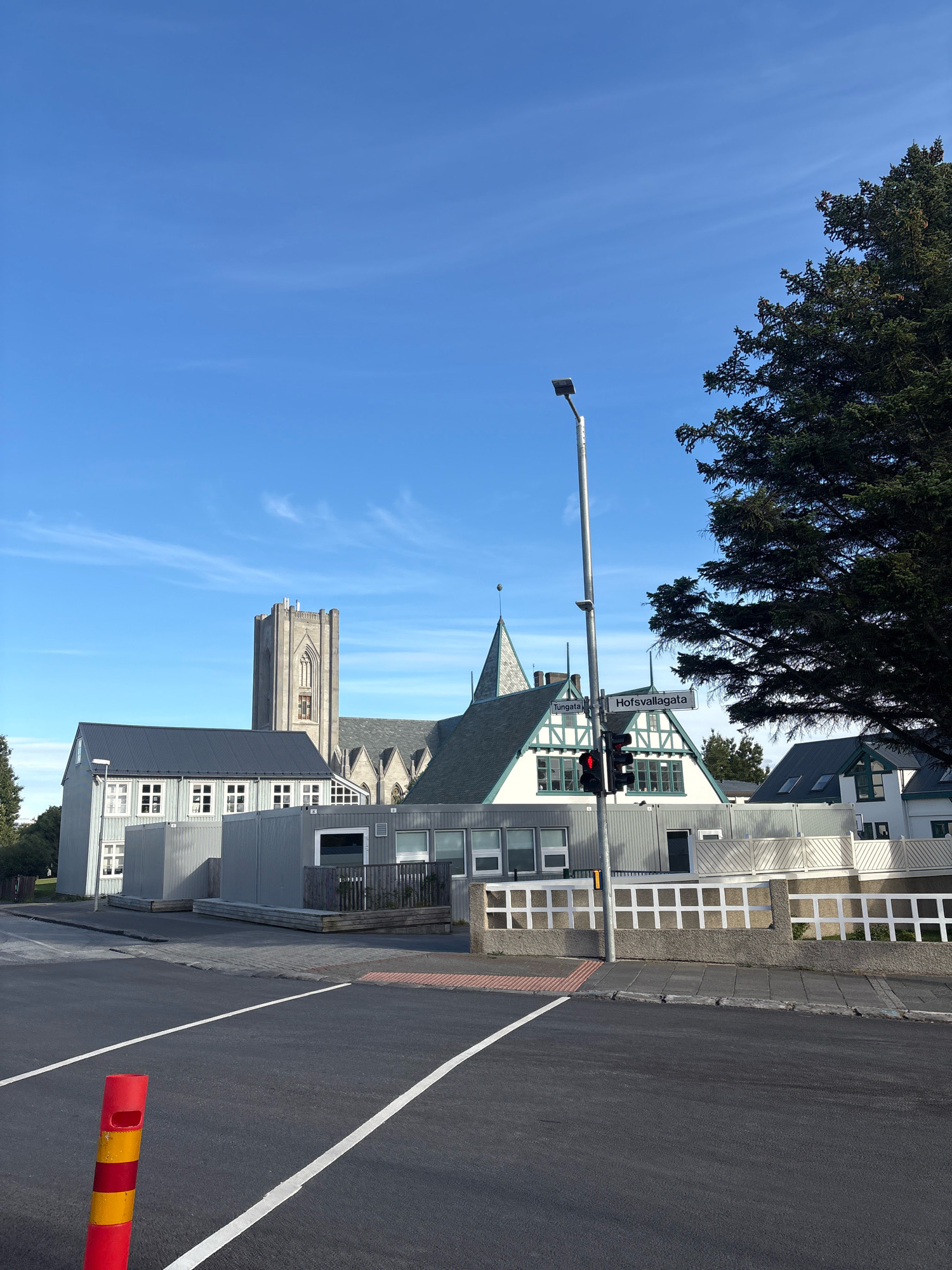 In Reykjavik, Iceland, a street view captures a bright blue sky with scattered wispy clouds. In the foreground, a street with white lane markings stretches across the frame. A red and yellow bollard stands on the left side of the road. A traffic light shows a red signal. To the right, a street sign indicates "Túngata" and "Hofsvallagata". Behind the sign, a building with a green roof and white trim is visible. Further back, a tall, grey stone church tower with Gothic architectural elements dominates the skyline. Several other buildings with grey and white exteriors and pitched roofs are also present. A large evergreen tree with dark green foliage stands on the right side of the image, partially obscuring the sky. The scene appears to be a daytime urban setting.