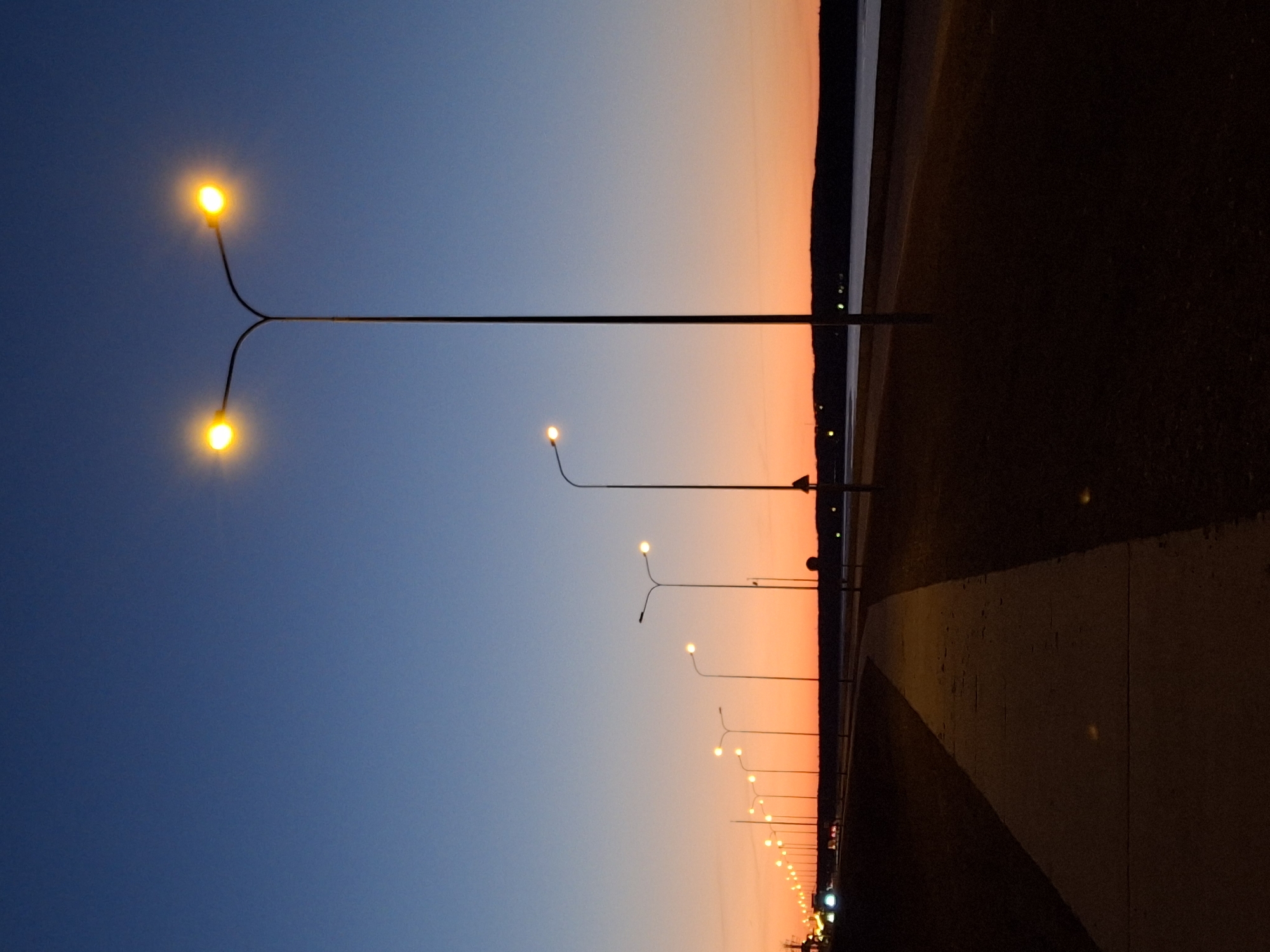 The photograph shows a row of streetlights along a road or promenade at dusk in the city of Chaikovsky, Russia. The sky is a gradient of blue to orange near the horizon. A body of water or land is visible on the right of the image. There is a walkway or road on the right side of the image. There are no people visible in the scene.