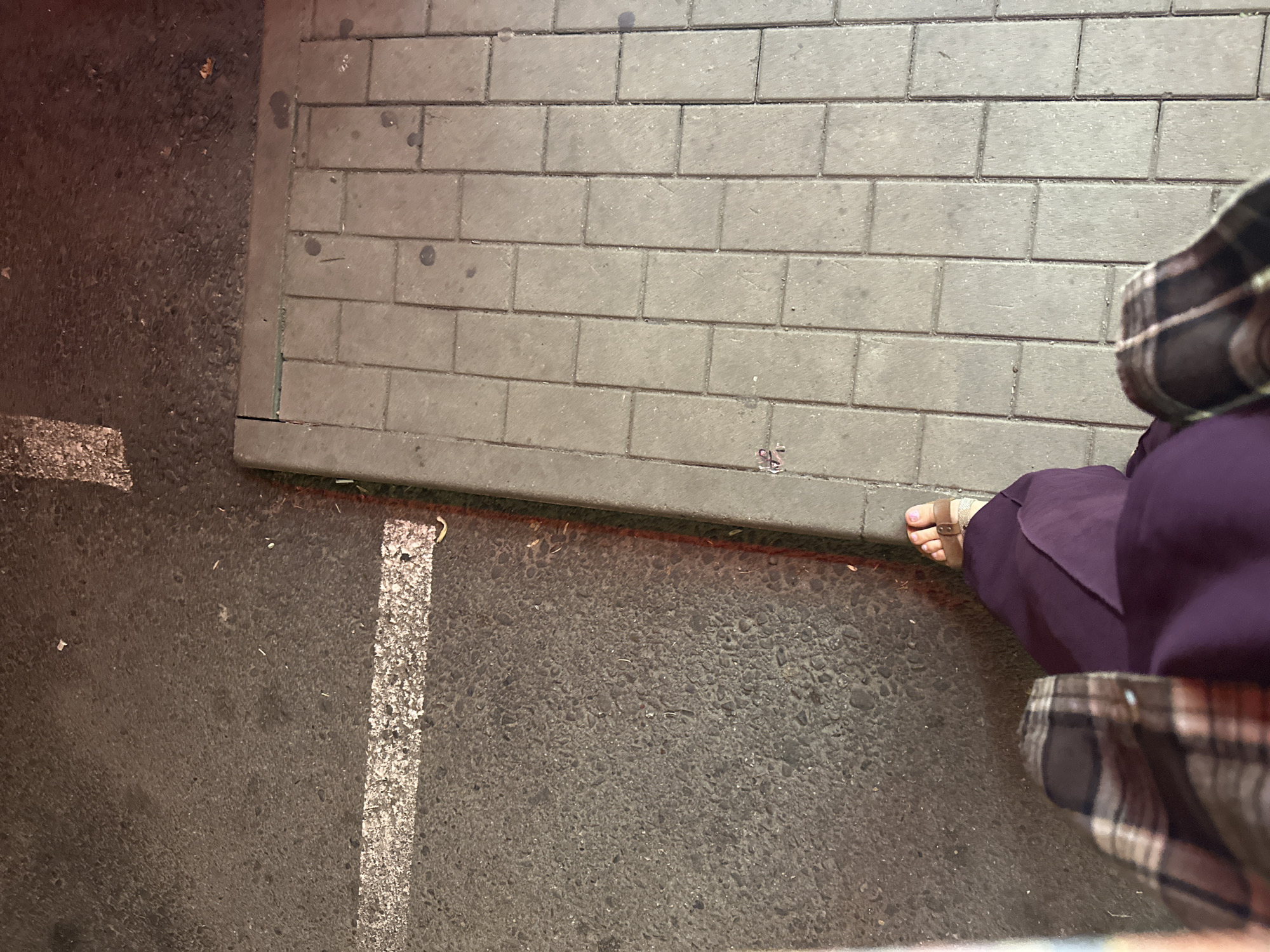 The image shows a person's feet and legs as they stand on a sidewalk next to a street. The person is wearing purple pants and a plaid shirt. One foot is visible, wearing a sandal. The sidewalk is paved with rectangular gray bricks, and the street is asphalt with a white line painted on it. The scene appears to be outdoors, likely in an urban environment. The lighting suggests it might be nighttime or dusk, with artificial lights illuminating the area. There are no other people or vehicles visible in the frame. The location is identified as Nevinnomyssk, Russia, though no specific landmarks or contextual clues in the image directly confirm this.