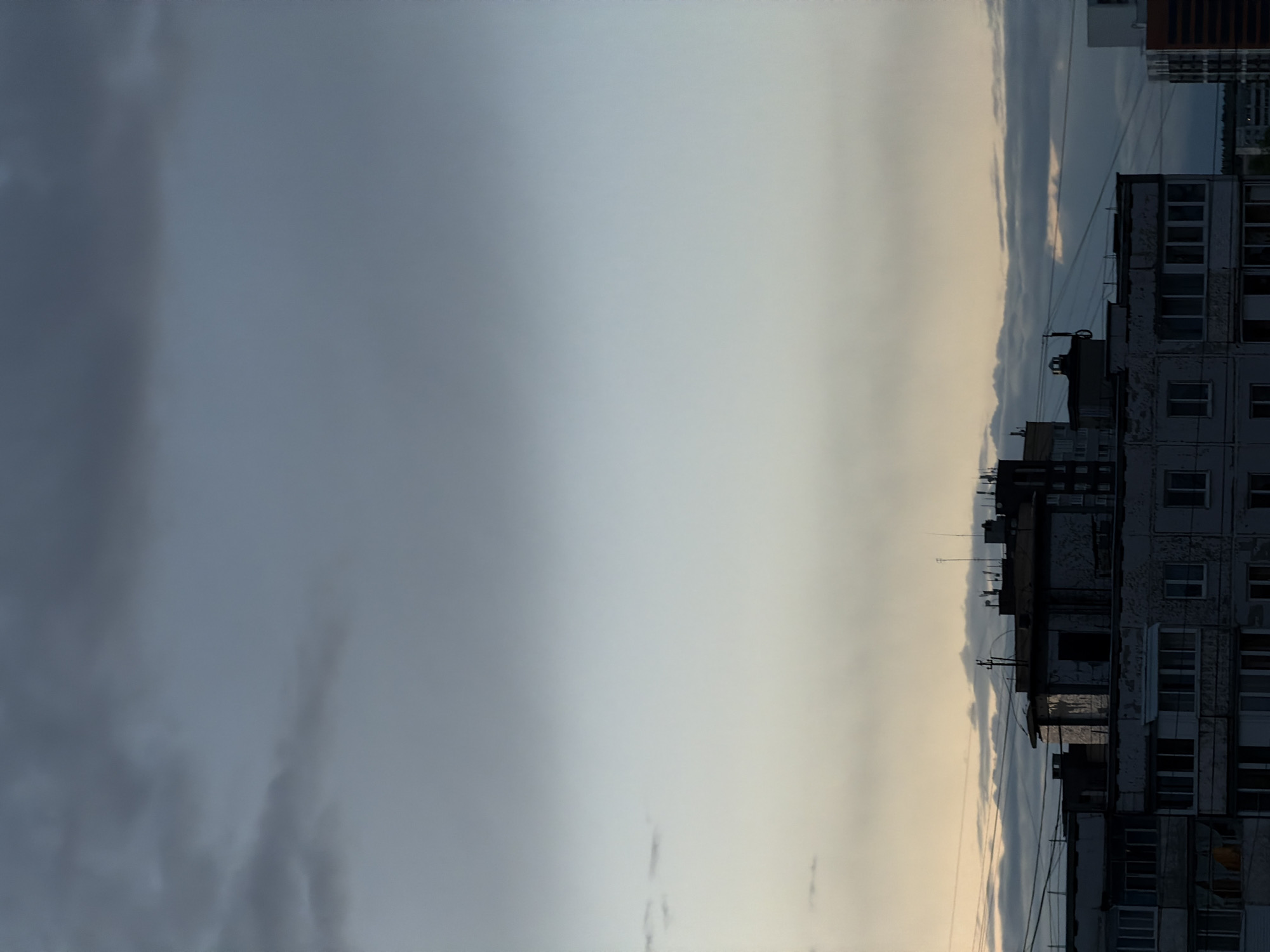 The image shows a cityscape in Perm, Russia. The right side of the image displays the facade of an apartment building with numerous windows. In the background are additional buildings and power lines. To the left, the sky transitions from a pale, cloudy gradient to a darker, overcast section, indicating possibly the beginning or end of a storm. The image is oriented vertically.