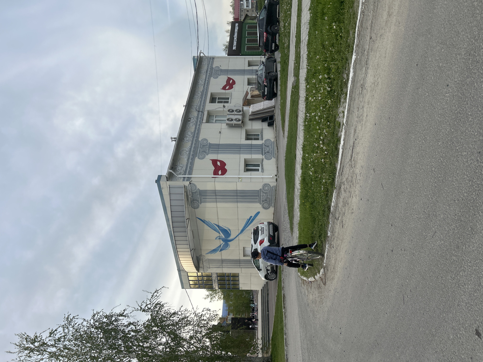 The image shows the exterior of a building in Uray, Russia, with a partial view of a street and sidewalk. The building has white walls adorned with painted grey columns, images of red masks on the upper levels, and a blue bird on the lower level. Several air conditioning units are mounted on the exterior wall. Two dark-colored SUVs and a light-colored car are parked alongside the building. A person on a bicycle is standing on the sidewalk in front of the building. The street appears to be paved and bordered by a strip of grass. The sky is overcast. A tree is partially visible in the foreground.