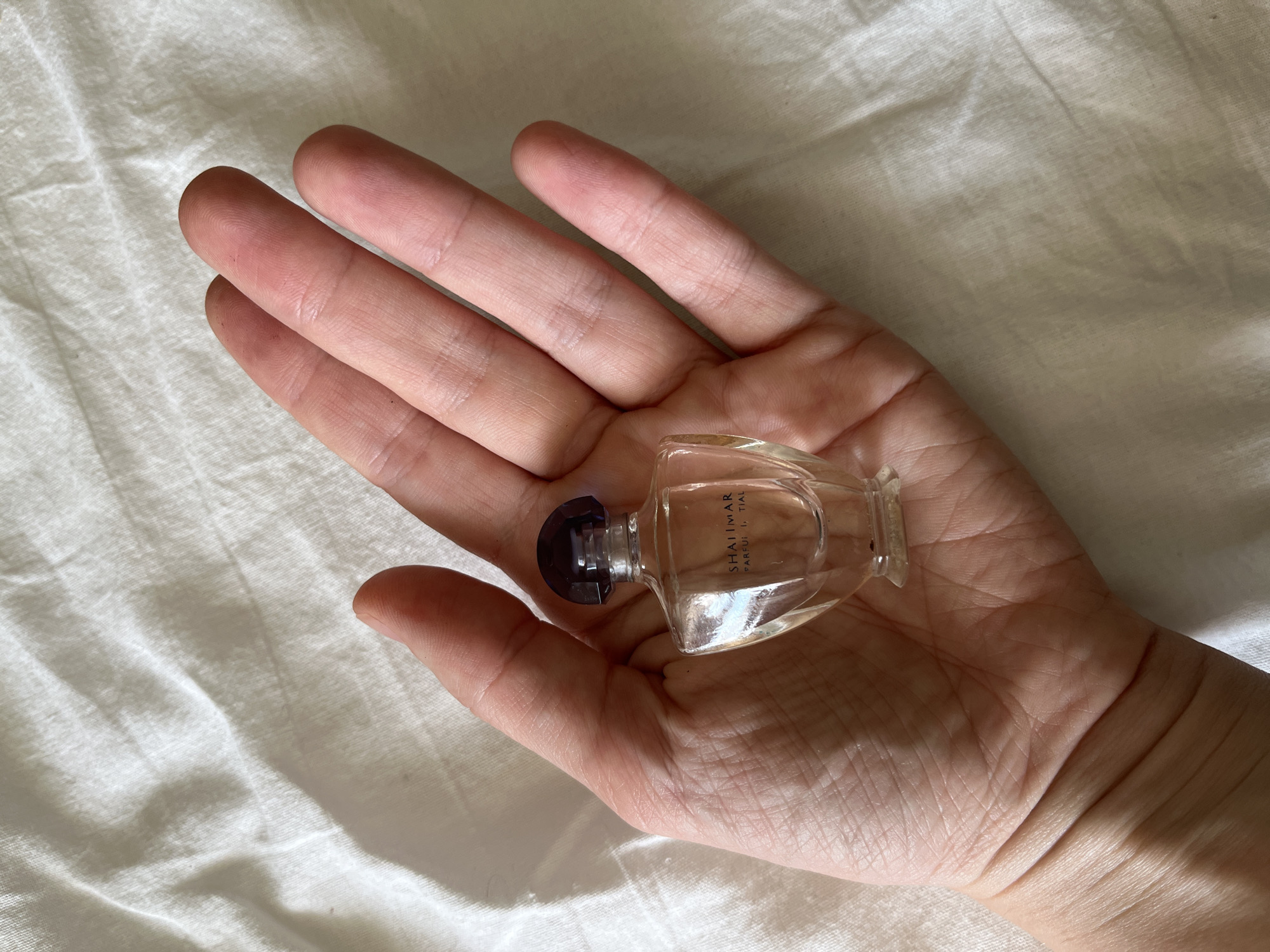 A hand is holding a small, clear glass perfume bottle with a dark blue cap. The bottle has text that appears to read "SHALIMAR PARFUM." The hand and bottle are resting on a white, textured surface, possibly a bedsheet. The image is well-lit, with natural light casting shadows. The background is out of focus, drawing attention to the main subject. There is no information in the image that can identify the location as Komsomolskoye, Ukraine.