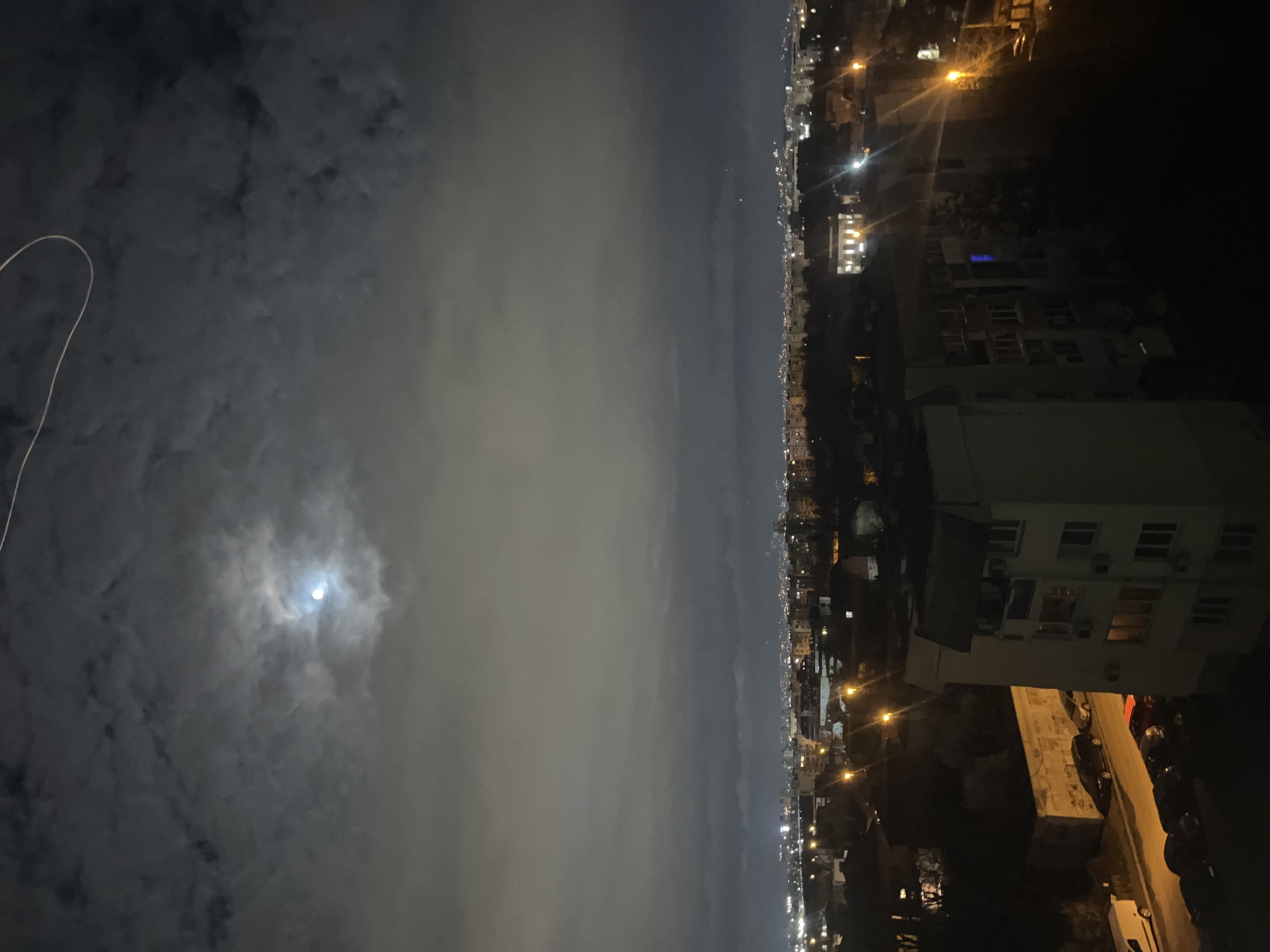 The image depicts a nighttime scene, likely from a high vantage point overlooking a city. On the left, the sky is predominantly covered with clouds, with a partially visible moon shining through. On the right, the city skyline is visible, characterized by numerous buildings, streetlights, and car lights. The perspective suggests the photo was taken from a building, looking out toward the city. The image might be in Sofia, Bulgaria. However, without any specific landmark that is visible in the image it is challenging to confirm that.