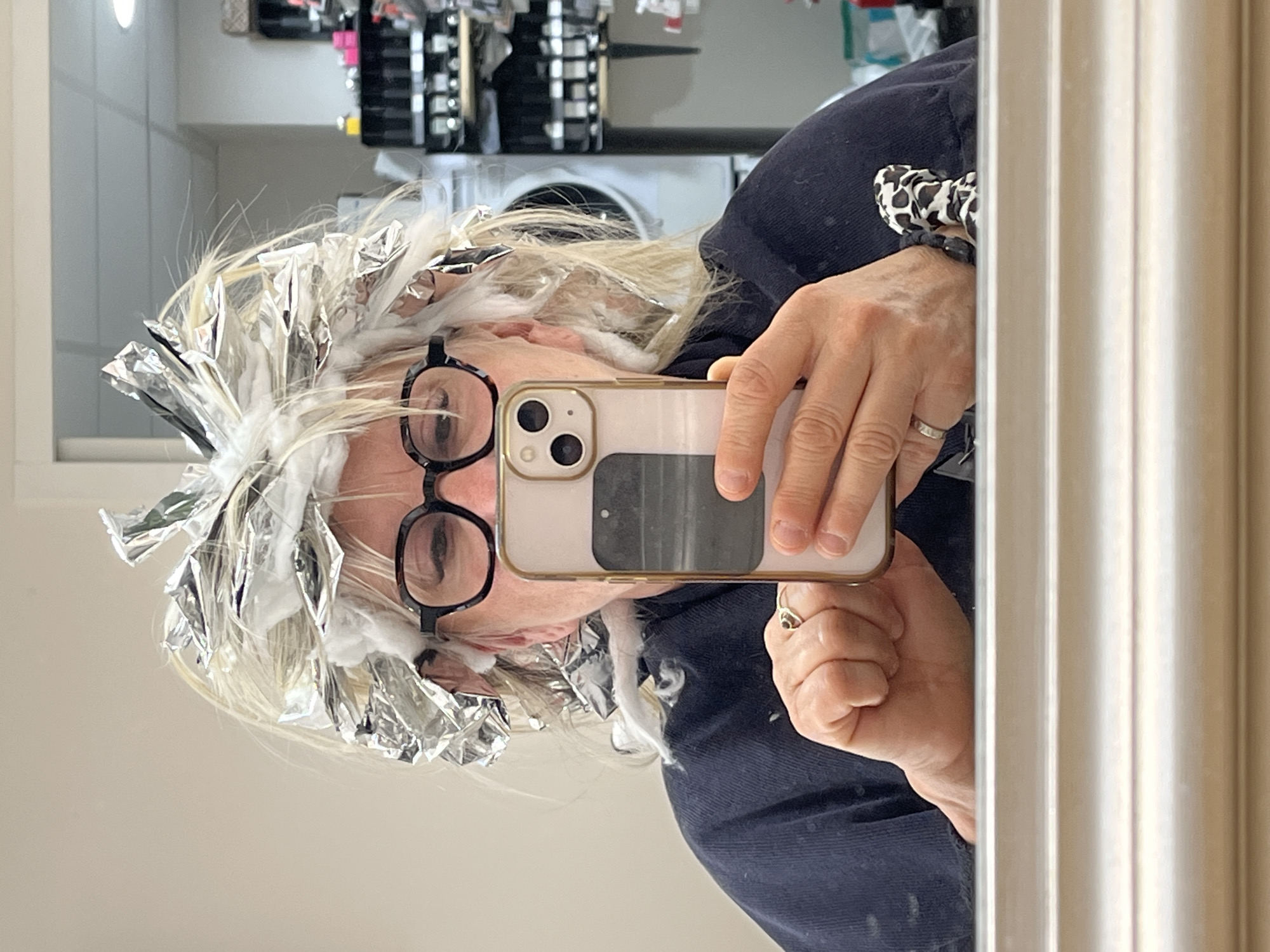 A woman with blonde hair and black-rimmed glasses is taking a selfie in a mirror. Her hair is partially covered with silver foil, suggesting a hair dyeing or highlighting process. She is holding an iPhone with a light-colored case. She is wearing a dark blue top and a patterned scrunchie on her wrist. The background includes a shelf unit with various items and a tiled wall. The environment appears to be a bathroom or salon-like setting. It is not possible to verify the location of the city of Montrond-les-Bains, France based on visual information.