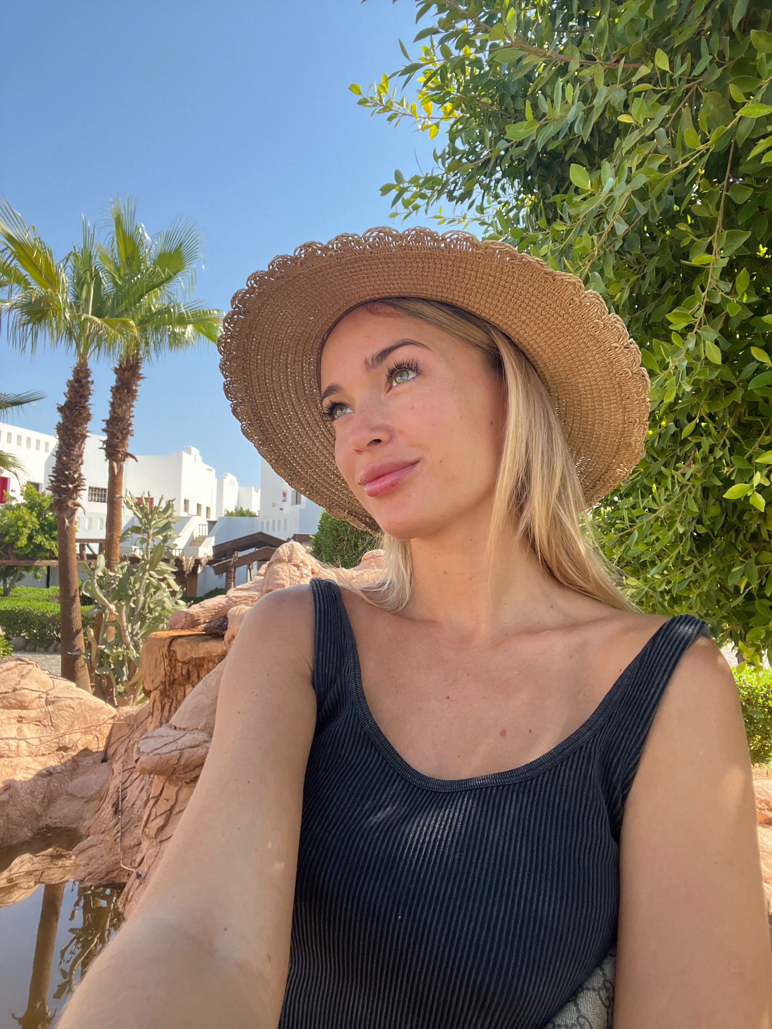 A young, blonde woman wearing a wide-brimmed straw hat and a dark ribbed tank top is captured in a close-up, selfie-style shot. She is looking upwards and to her right, with a slight smile on her face. Her eyes are light green, and she has a smattering of freckles across her nose and cheeks. The background features a bright blue sky, lush green foliage, palm trees, and white buildings with arched windows, indicative of resort architecture. In the foreground, there are textured, reddish-brown rock formations and a body of water, likely a pool or pond. The overall scene suggests a sunny, outdoor location in Sharm El Sheikh, Egypt, possibly a hotel or resort area.
