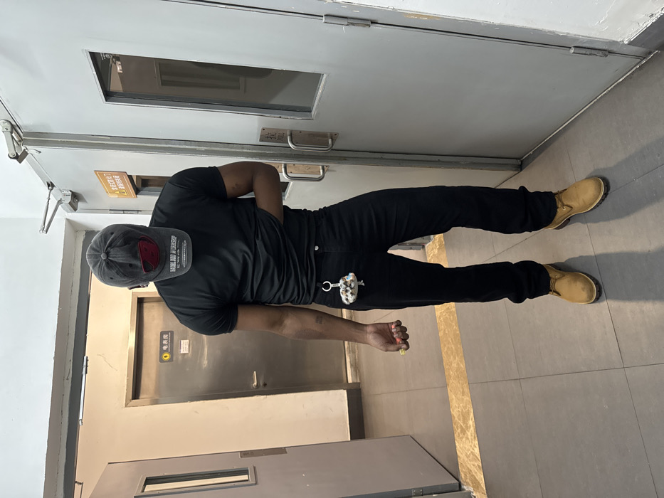 A single individual stands upright in an indoor corridor within Yuzhong District, China. The person, an adult male with a dark complexion, is facing slightly towards the left of the frame. The individual wears a black short-sleeved t-shirt, black trousers, and light brown work boots. A dark grey baseball cap, featuring a red design and the text "LABEL AND WORKSHOP", is pulled low, completely obscuring the face. A tattoo is visible on the inner aspect of the left forearm.
The individual's left hand is lowered, holding a small, colorful object, while the right hand is tucked beneath the front hem of the t-shirt, near the waistline. A multi-colored, beaded keychain is attached to a belt loop on the left side of the trousers.
The environment consists of a grey tiled floor, bisected by a horizontal stripe of light brown, marble-like material. The walls are light-colored. Three metallic doors are visible: a partially open light grey door on the far left, a closed dark grey door in the center-background with a yellow sign indicating "电表房" (Electric Meter Room), and a closed light grey door on the right with dual horizontal handles and a red sign above it, partially visible, which reads "安全出口" (Emergency Exit) and "防火门" (Fire Door). A door closer mechanism is mounted above the rightmost door. Overhead lighting illuminates the scene, casting shadows.