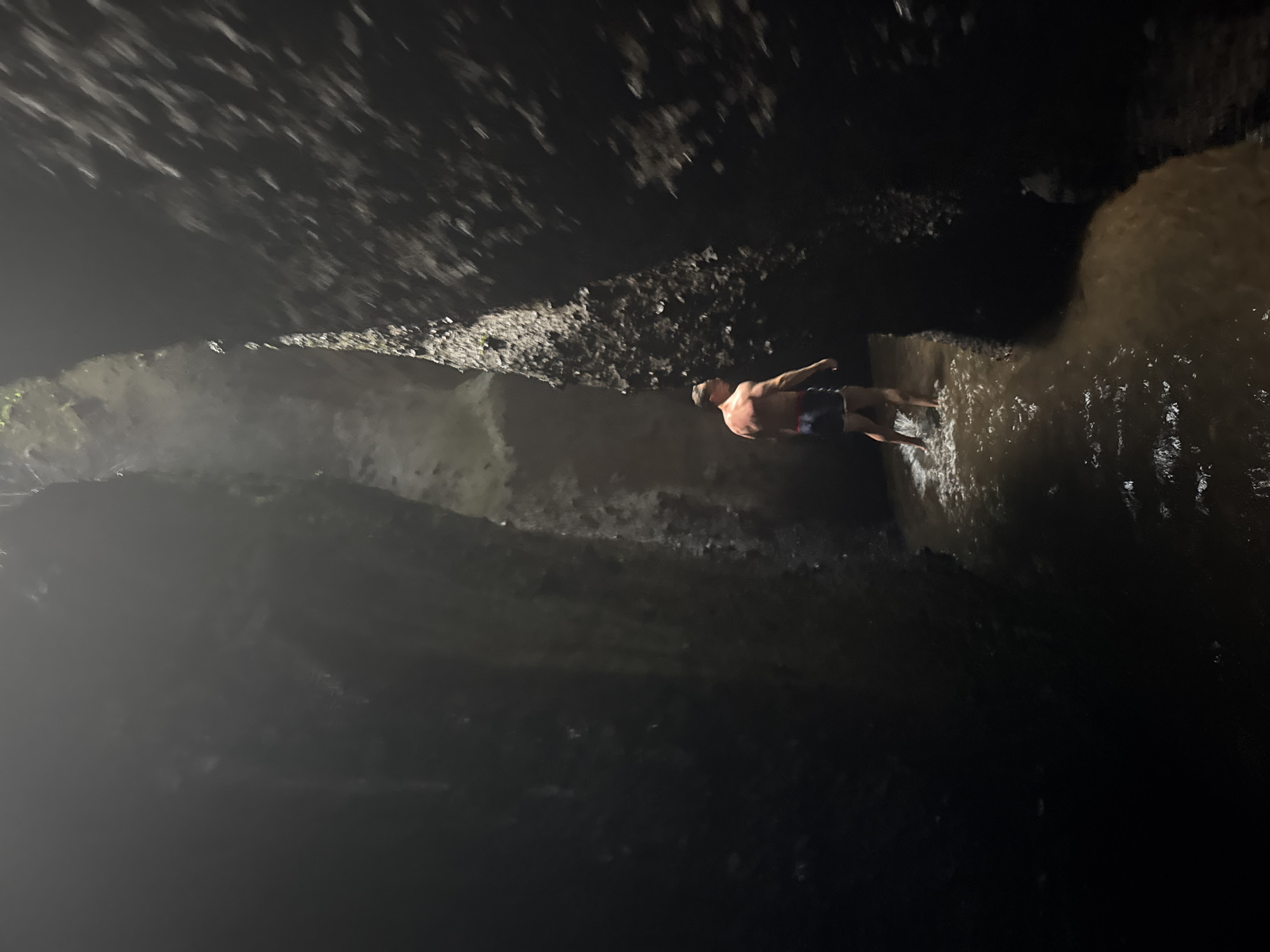 A bare-chested man wearing swim trunks walks through a narrow, dark gorge with water flowing around his ankles. The walls of the gorge are rough and uneven, with some vegetation visible on the upper edges. A shaft of light penetrates from above, illuminating the man and the water. This scene likely depicts Tukad Cepung Waterfall in Bangli, Indonesia, a popular tourist destination known for its unique cave-like waterfall.