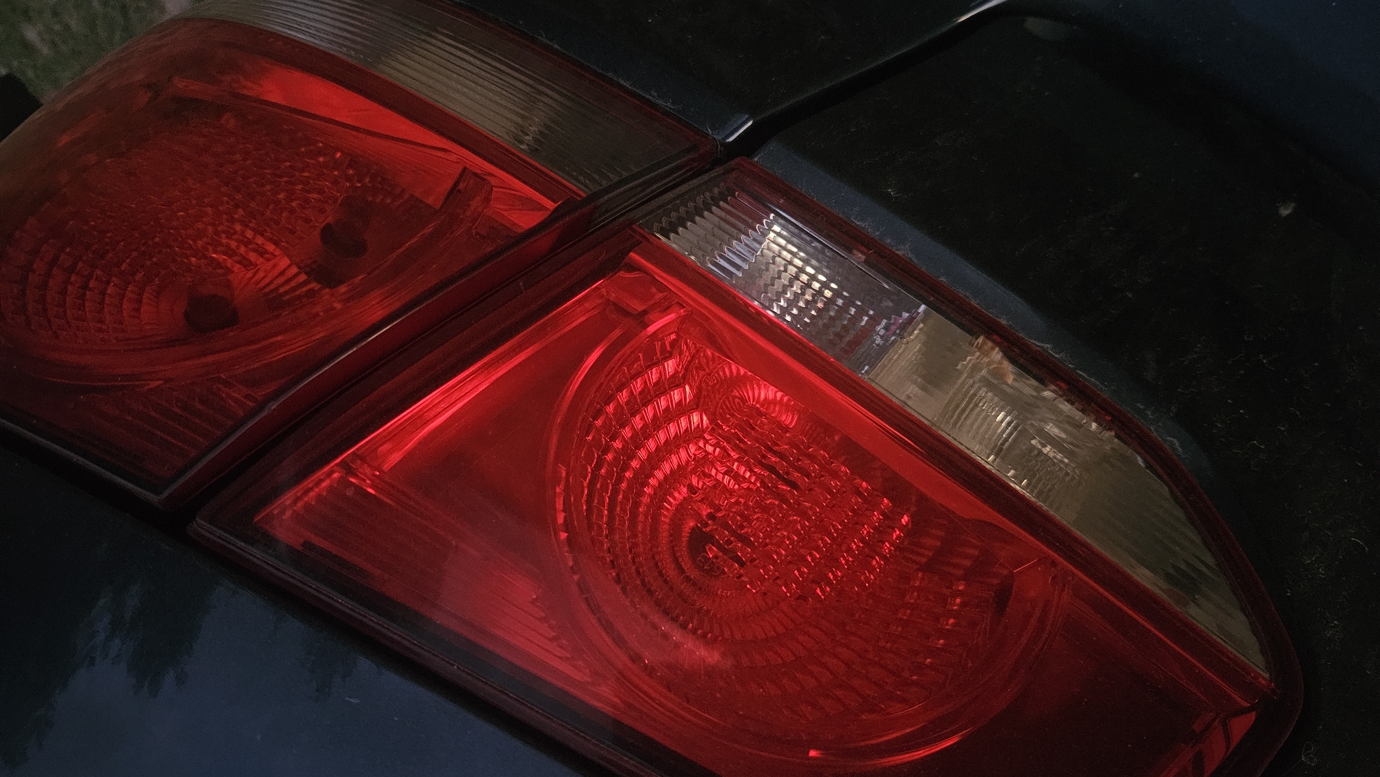 The image shows a close-up, angled view of a car's rear taillight assembly. The taillight is illuminated with a bright red light, revealing a textured, circular pattern within the red lens. Adjacent to the red section, there is a clear or lightly tinted lens with a ribbed or grooved surface. The car's body, which appears to be dark in color, is visible surrounding the taillight assembly. The lighting suggests it is either dusk or night, and the scene is a close-up focus on the automotive component. No location in the city of Unknown, Unknown can be determined from the image.