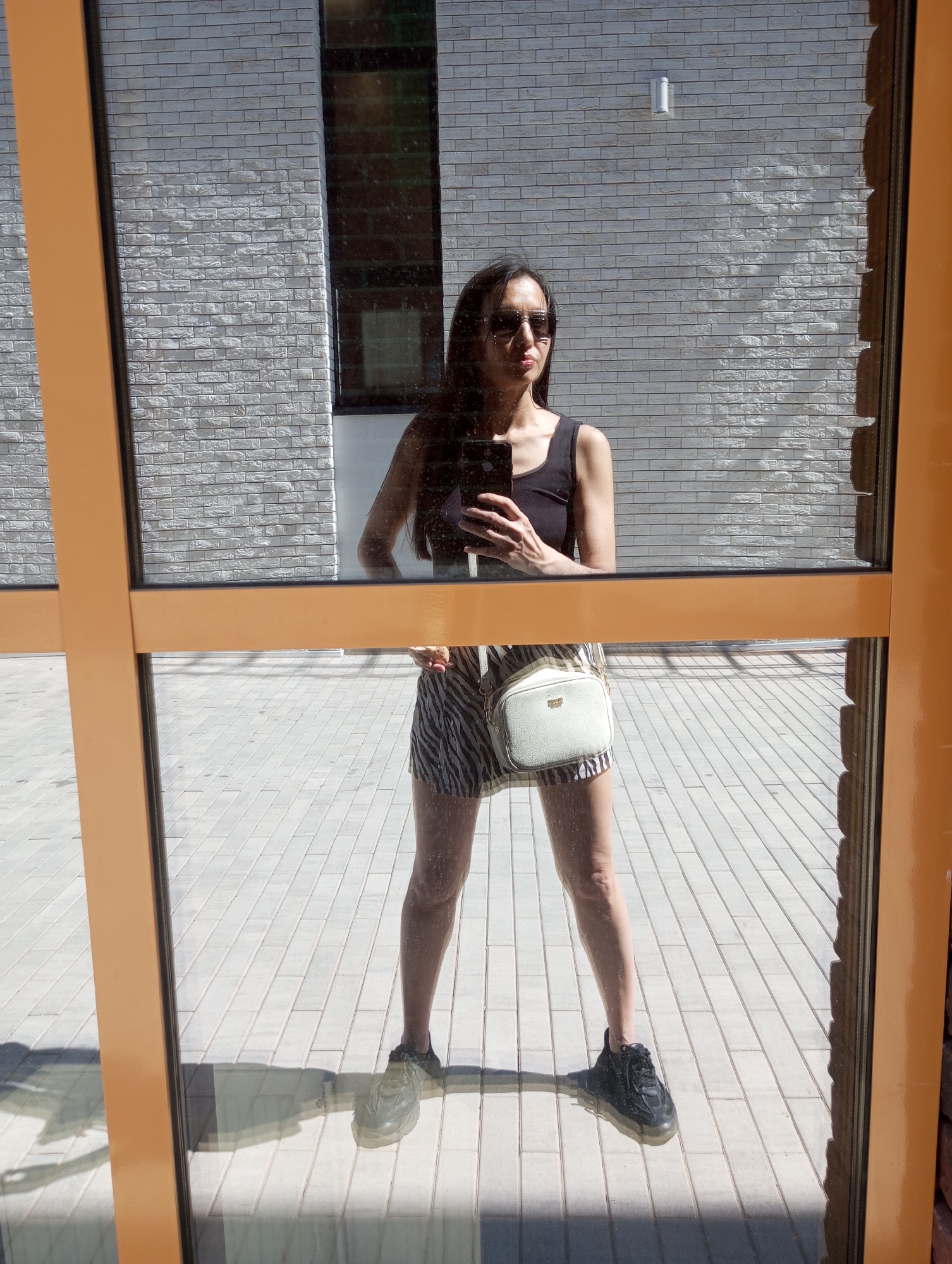 A woman stands reflected in a large window, holding a smartphone. She wears sunglasses, a black tank top, a zebra-striped skirt, and black sneakers. A white crossbody bag hangs on her shoulder. The window has a brown frame and reflects the surrounding environment, including the ground paved with rectangular tiles and a brick building in the background. The scene is set in Pervomaysky, Russia.