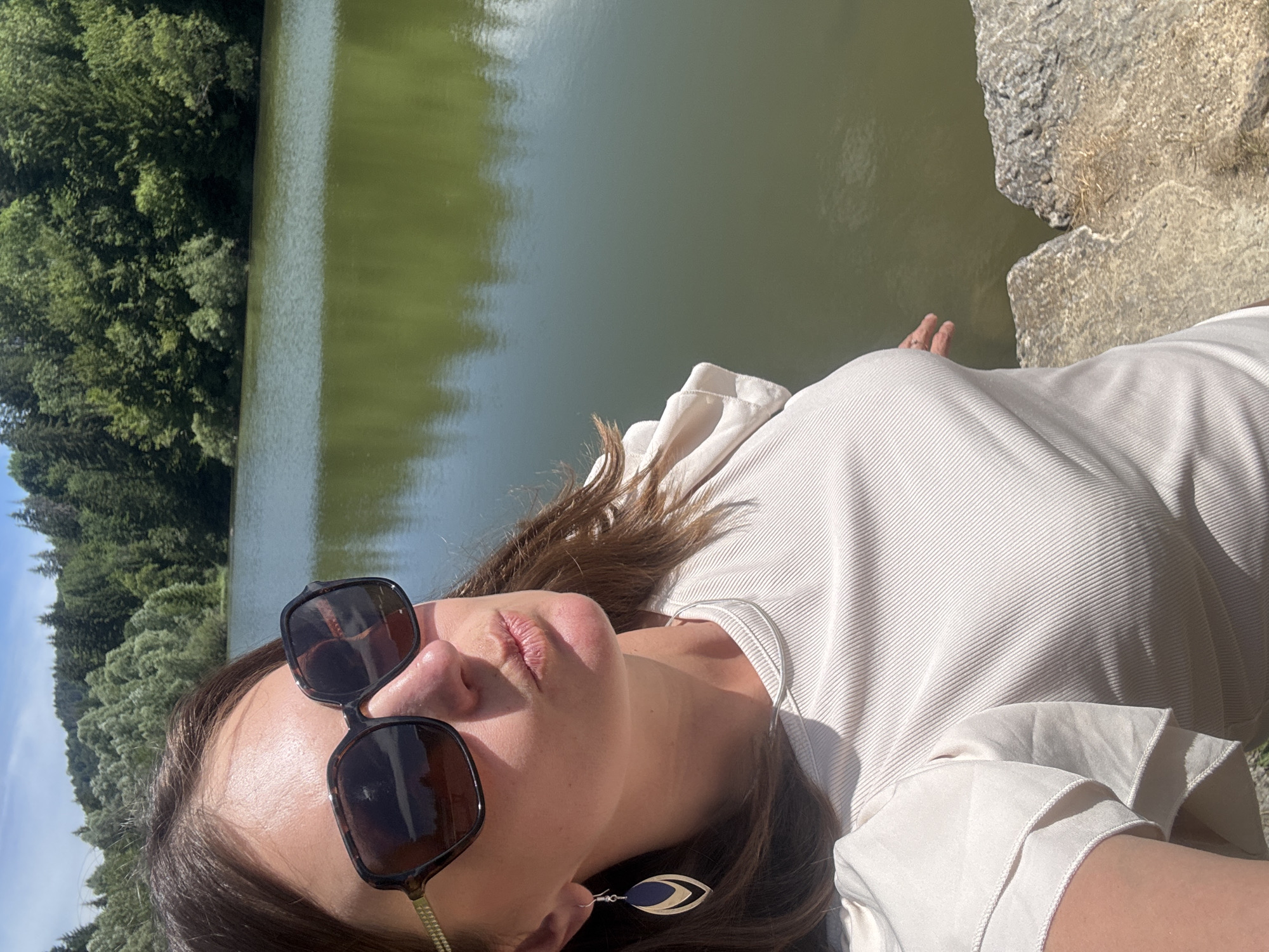 A medium shot captures a woman taking a selfie on a sunny day with a body of water and a treeline in the background, likely in Coteaux du Lizon, France. The woman, facing the camera, wears large, dark sunglasses, a light-colored ribbed top with ruffled sleeves, and dangling earrings. She is positioned on a rocky outcrop, with her left arm visible and her right hand reaching towards the water. The calm, green water reflects the sky and the dense, lush green trees that line the opposite bank. The sky is a clear, bright blue with some wispy clouds.