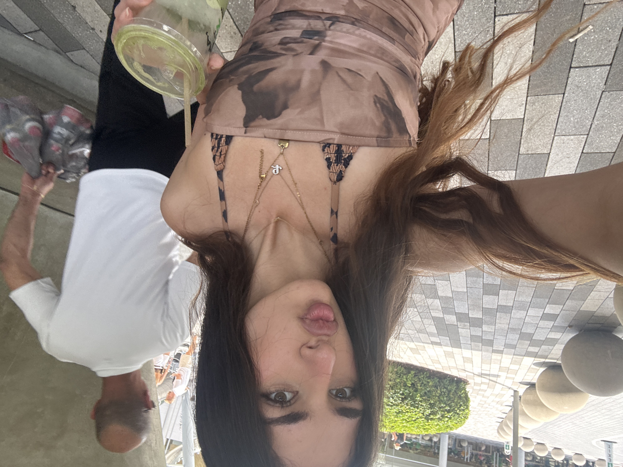 The image depicts a woman taking a selfie. She has long brown hair and is wearing a brown patterned dress with straps. She is also wearing gold necklaces. She is holding a drink with a straw in her left hand. In the background, a man is carrying bags. The location appears to be an outdoor shopping area with tile flooring, shrubs, and spherical outdoor lights. Based on the lighting and architectural style, it appears to be located in the city of Aventura, United States of America.