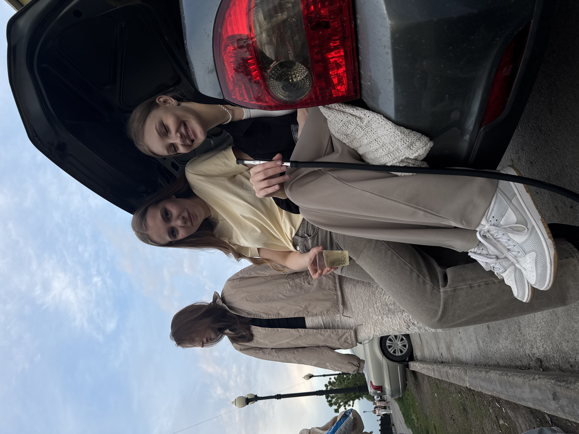 Three young women are sitting in the open trunk of a dark-colored car, possibly in the city of Perm, Russia. The car is parked on the side of a street with street lamps visible in the background. One woman is holding what appears to be a hookah pipe, while another is holding a clear, rectangular object. The women are casually dressed. The trunk lid is open, revealing the car's taillight. The sky is partially visible. Other parked cars and pedestrians can be seen in the background.
