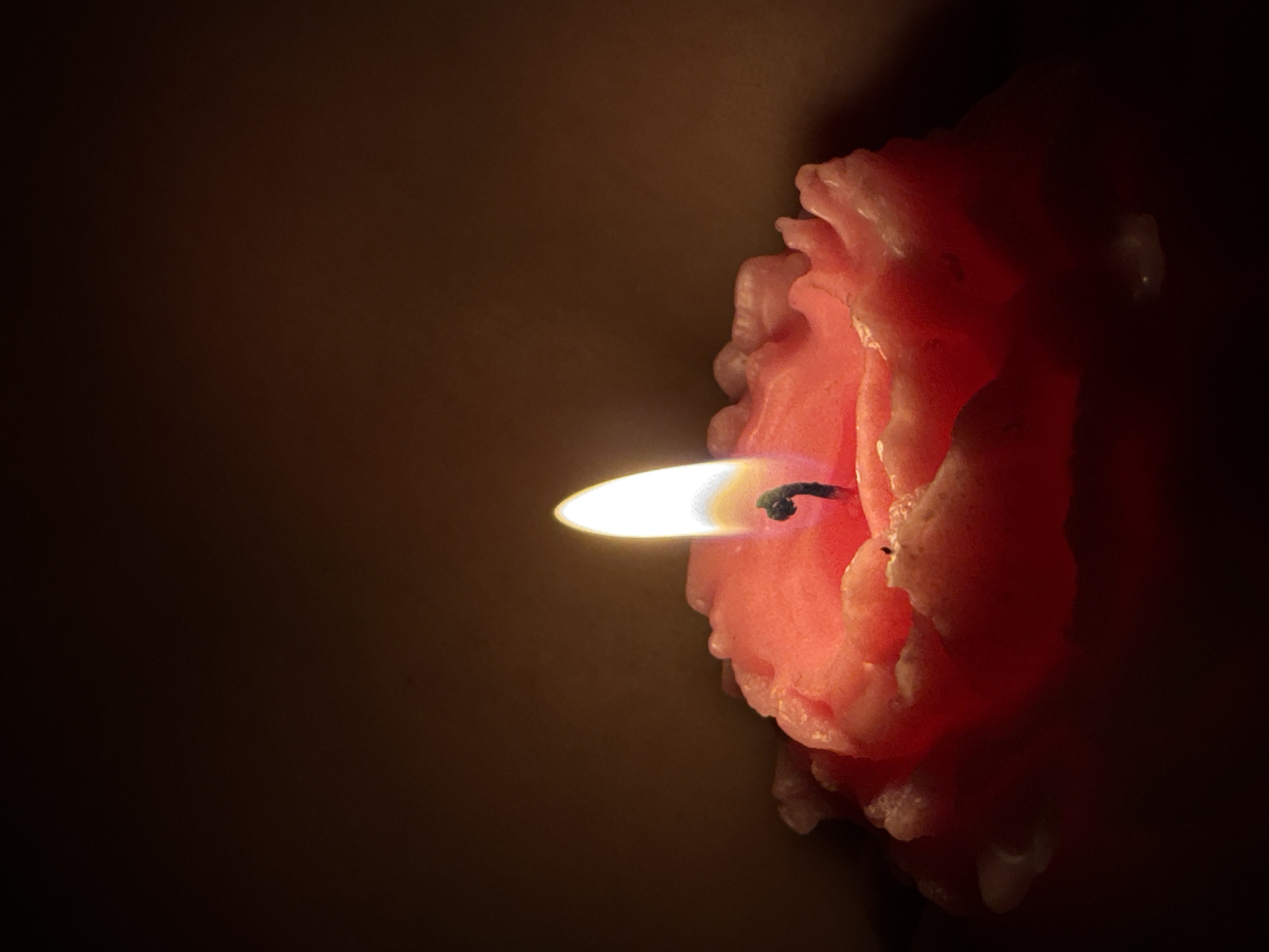 The image is a close-up shot of a lit pink candle with a ruffled edge, set against a dark, blurred background. The flame of the candle is bright white at the core, transitioning to yellow and then a faint orange at the tips, casting a warm glow. The wax of the candle has melted unevenly, creating a textured, petal-like appearance around the wick. The scene is dimly lit, with the candle being the primary light source, suggesting an indoor setting possibly at night. There are no people or other objects visible in the frame. The image does not provide any information about the city of Chaikovsky, Russia.