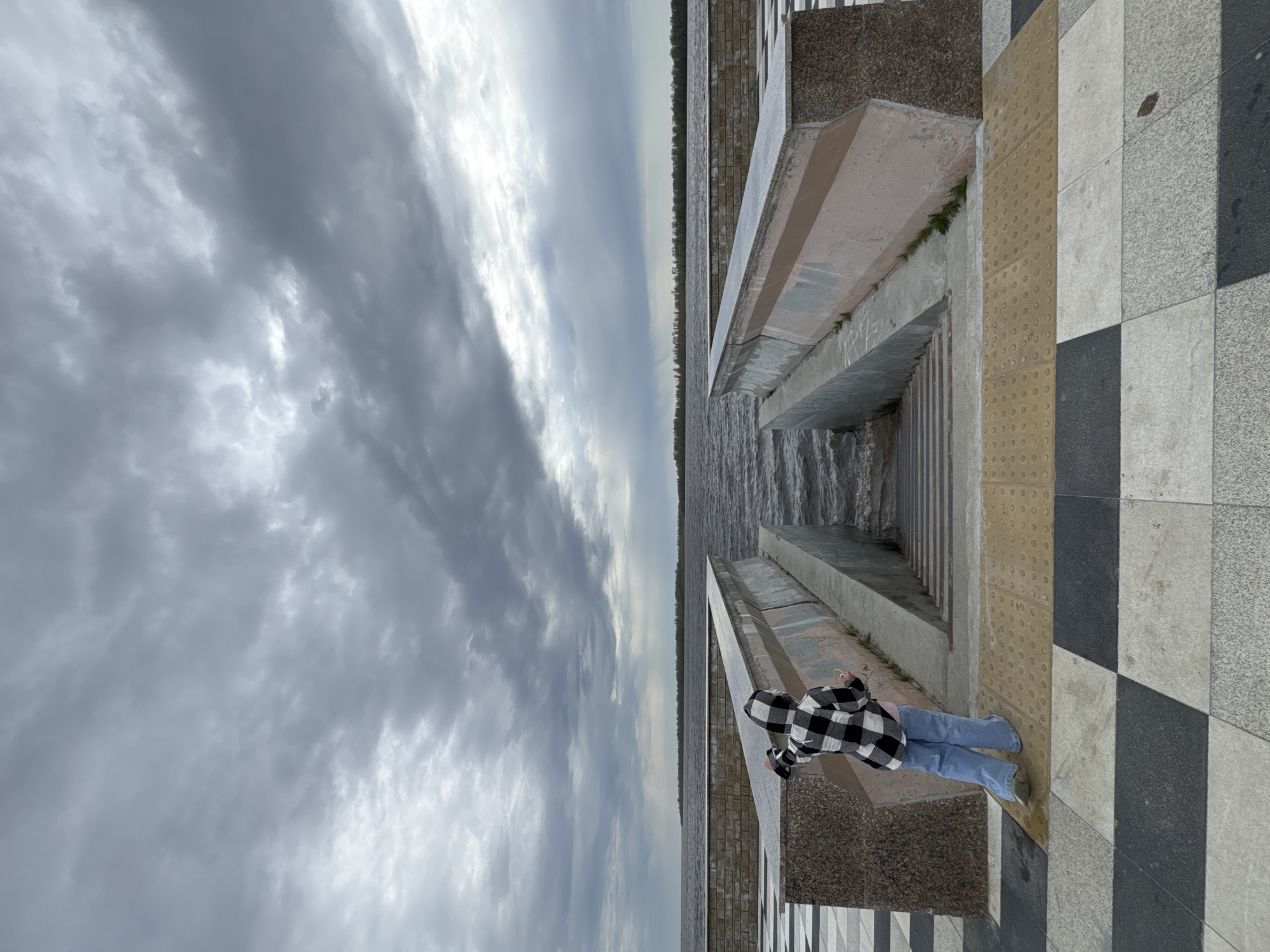 A person stands on a checkered pavement next to a stone structure, with a body of water and cloudy sky visible in the background. The scene is set in Nizhnevartovsk, Russia. The person is wearing a black and white checkered jacket and jeans. The stone structure is made of concrete and brick. The body of water is dark and choppy. The sky is overcast with grey clouds.