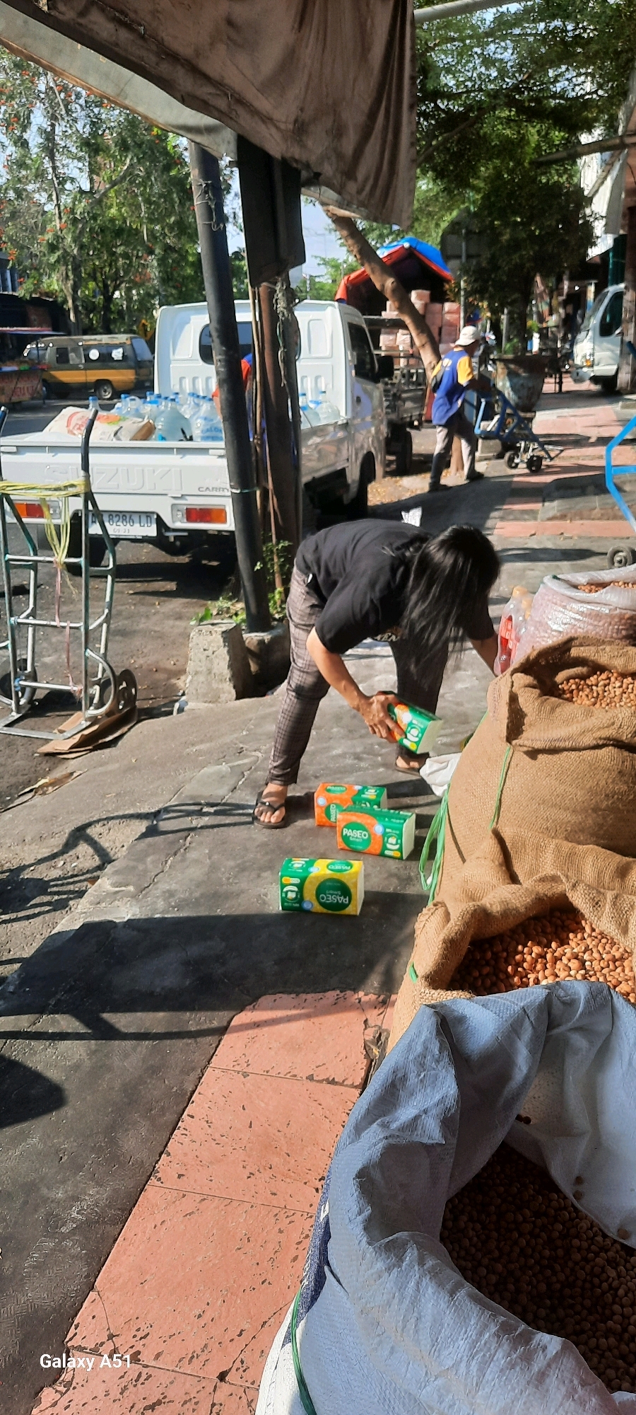 A low-angle, close-up shot captures a person bending down on a paved sidewalk in Beru, Indonesia, interacting with packages of tissues. The individual, wearing a black t-shirt and checkered pants, is in the process of placing or arranging three packages of "Paseo" brand tissues on the ground. Two of these packages are bright green and orange, while the third is yellow and green. The sidewalk is a mix of gray concrete and reddish-orange tiles. In the background, a white Suzuki Carry truck is parked, loaded with several large plastic bottles of water and sacks of what appears to be goods. To the left of the truck, a metal handcart stands on the sidewalk. Further in the background, trees and other vehicles are visible, suggesting a street or market environment. The lighting indicates it is daytime, with harsh shadows cast by the overhead structure and other objects.