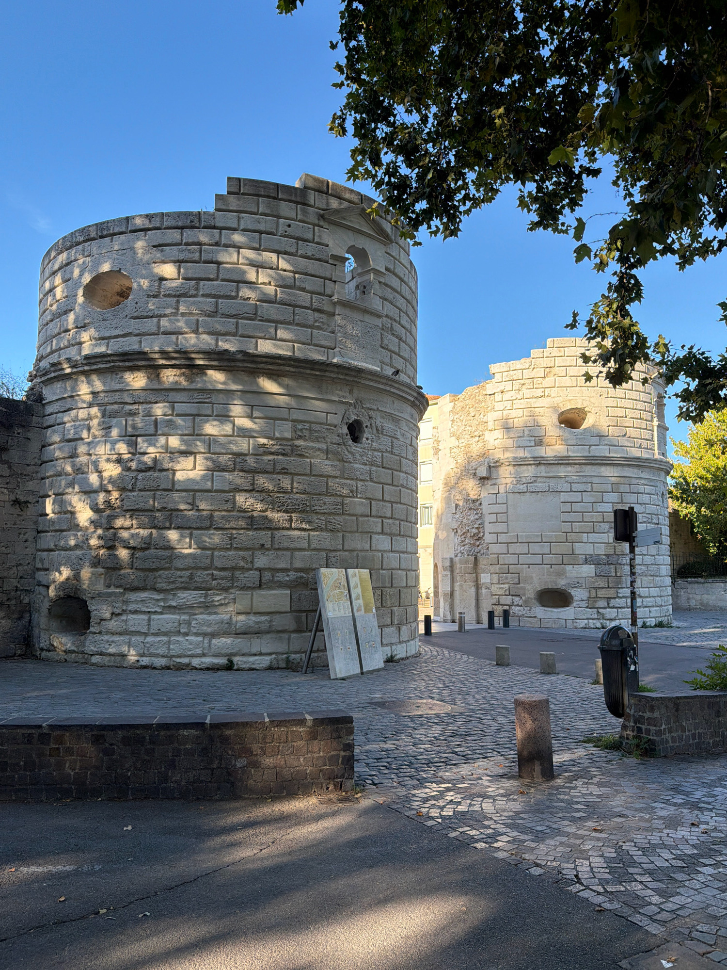 The image shows the Porte de la Monnaie in Arles, France, on a clear day. The ancient Roman gate, constructed of large, pale stone blocks, features two cylindrical towers. The tower on the left is more intact, with a visible circular opening and a crenellated top. The tower on the right is partially ruined, with sections of its stone facade missing. The ground in front of the towers is paved with cobblestones, and a paved road runs between them. Several small, cylindrical bollards are placed along the road. To the right, a black trash can and a low, dark brick wall are visible. Information panels are leaning against the left tower. The scene is framed by the dappled shadow of tree leaves from a large tree in the upper right corner. The overall impression is one of historical preservation within a contemporary urban setting.