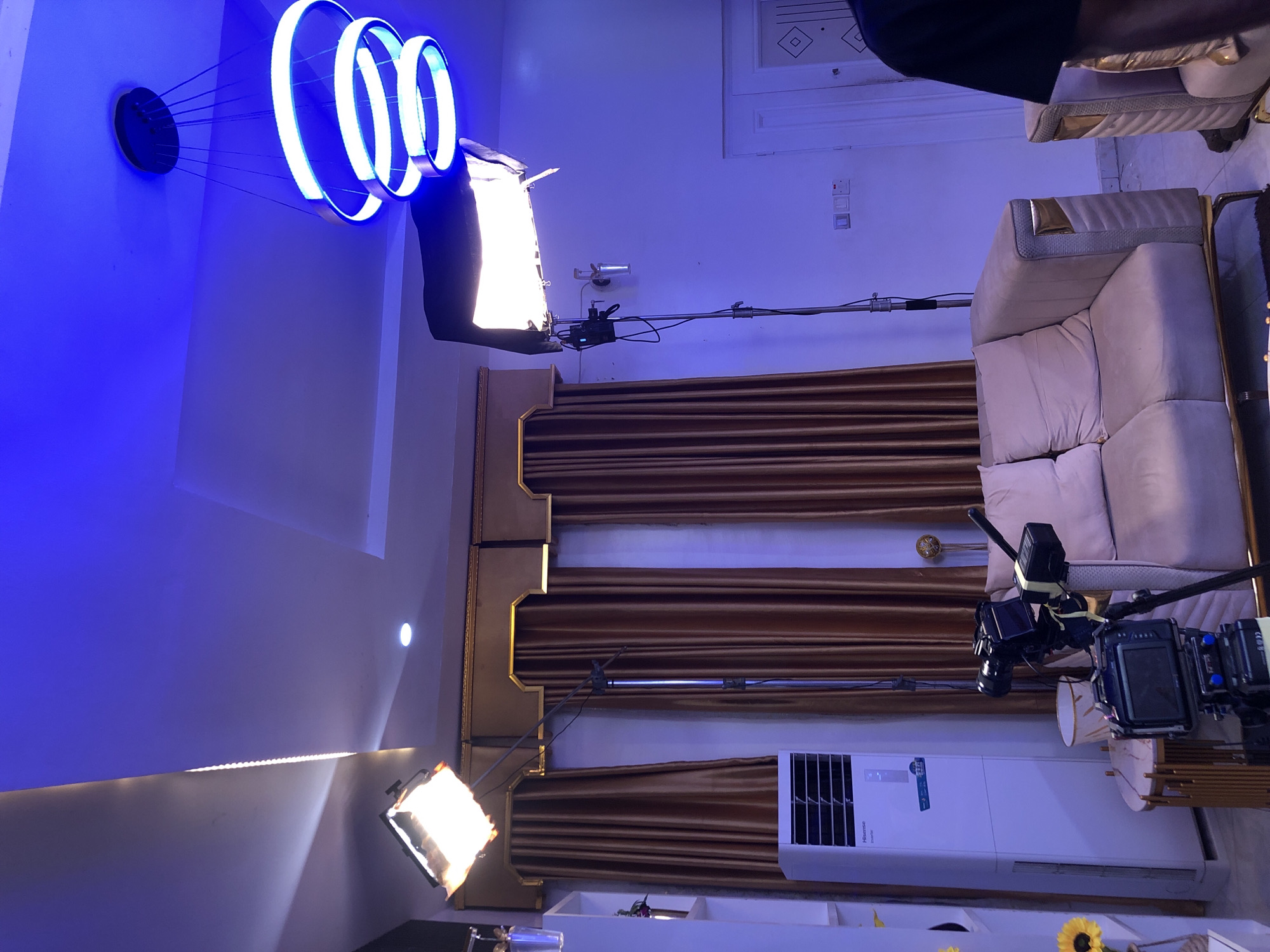 This image captures an indoor setting, likely a living room or a dedicated studio space, arranged for a photoshoot or video production. The walls are painted a light blue or purple, heavily influenced by the artificial lighting.
Prominent objects include various professional lighting and camera equipment. On the left, three concentric circular lights with glowing blue rims hang from the ceiling, casting a blue hue across that section of the room. Below these, a large, rectangular LED panel light with a diffuser emits intense white light, angled upwards. Another similar LED panel light is visible towards the bottom right, also emitting bright white light. A long boom arm or stand extends horizontally across the middle of the frame, supporting lighting equipment.
On the right side of the image, a cream or off-white sofa with textured upholstery and gold accents provides a seating area. Next to it, a small, round table with a marble-like top holds what appears to be camera equipment, including a camera body and attached monitors or recording devices. Further right, another camera is visible on a stand or tripod.
The background features dark brown curtains with intricate gold trim, adding a touch of elegance to the setting. Below the curtains, a white "Hisense inverter" air conditioner unit is mounted on the wall. Below the air conditioner, a shelf holds decorative items, including a vase with what appear to be sunflowers. Power outlets and light switches are visible on the far wall.
The scene suggests a professional setup, with no people explicitly visible but their presence implied by the complex arrangement of equipment. All illumination comes from artificial light sources, making the time of day undeterminable.