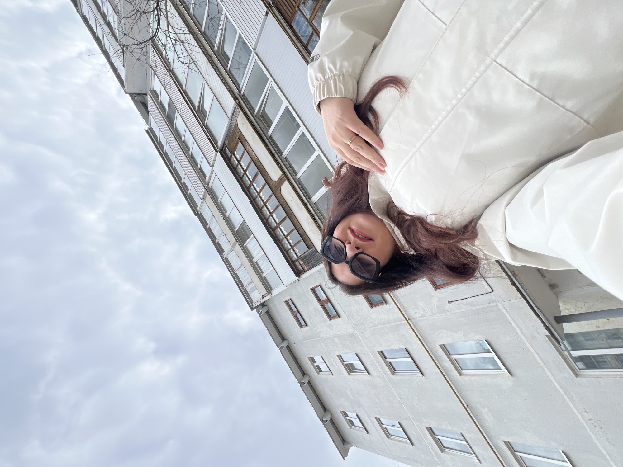 This selfie-style portrait captures a woman positioned against a backdrop of urban buildings and an overcast sky in Noviy, Russia. The woman, likely the photographer, occupies the foreground, looking directly at the camera. She has long, dark reddish-brown hair and wears large, dark sunglasses. Her attire includes a light-colored, possibly cream or beige, jacket. Her left hand is visible near her hair, adorned with a simple ring on her ring finger. Her lips are slightly parted, revealing a subtle hint of red lipstick, suggesting a relaxed expression.
The background features two multi-story buildings, viewed from a low angle, giving a sense of looking upwards. The taller building on the left exhibits several balconies and numerous windows, some with white paneling and dark window frames. A second building, a grey concrete structure with scattered rectangular windows, is partially visible on the right. Bare tree branches extend into the upper left of the frame, hinting at either winter or early spring. The sky is uniformly overcast with soft, grey clouds, indicating a cloudy daytime. No visible text is present.