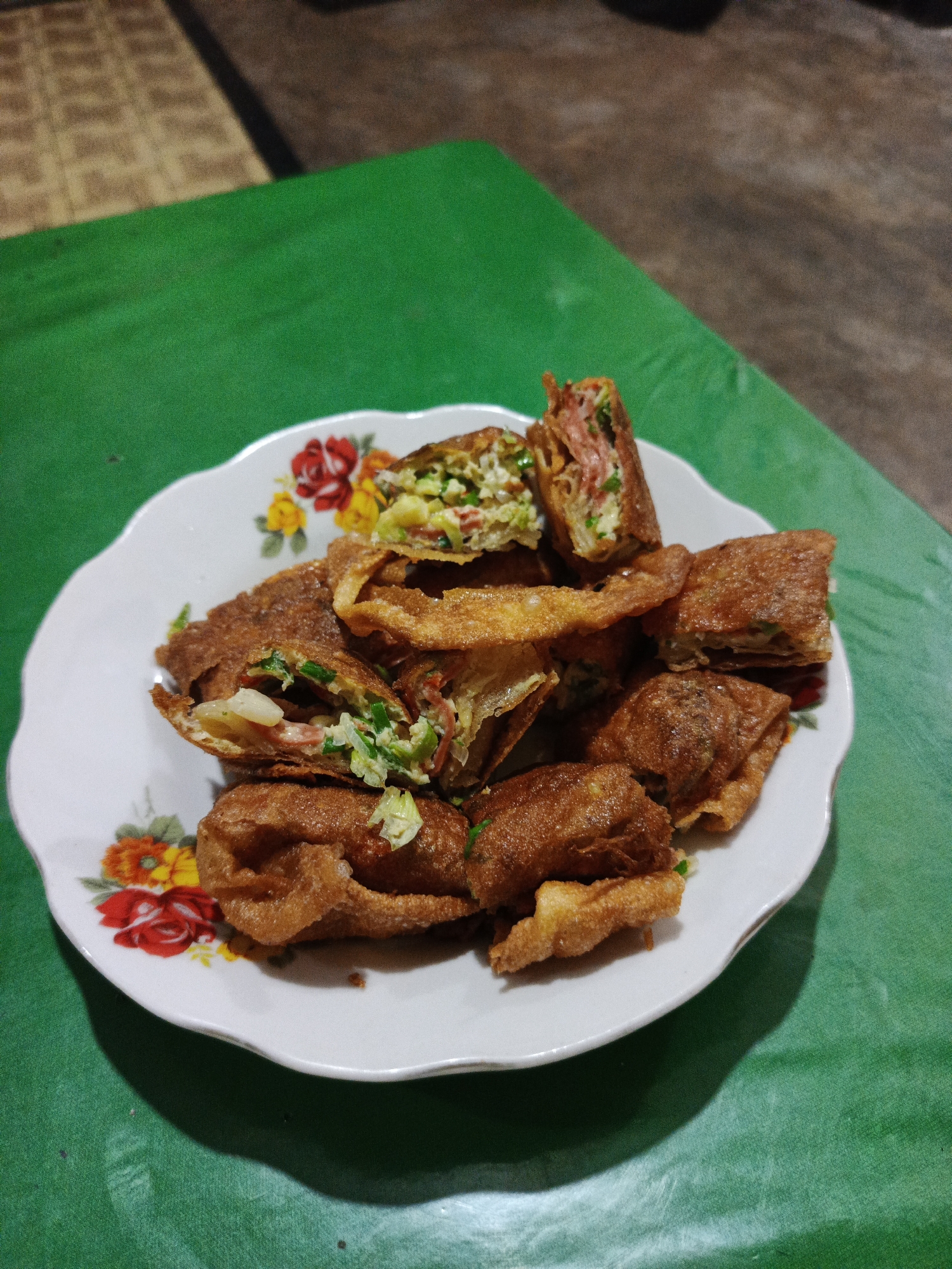 A white, floral-patterned plate filled with fried savory pastries sits on a green surface. The pastries are golden brown and appear to be stuffed with a mixture of meat and vegetables. Some of the pastries are whole, while others have been cut in half, revealing the filling. The plate has a scalloped edge and is decorated with red, yellow, and white flowers. The green surface beneath the plate has a textured, slightly wrinkled appearance. In the background, a blurred, woven mat and a darker, out-of-focus area suggest an indoor or covered outdoor setting. The lighting appears to be from above and slightly to the left, casting a shadow of the plate onto the green surface. The overall scene is simple and focuses on the food.