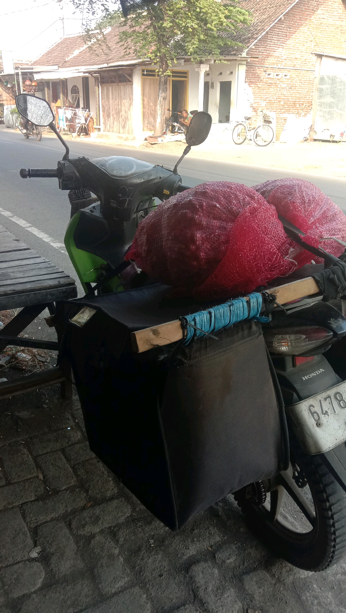 In Karanganyar, Indonesia, a parked motorcycle is loaded with two large, red net bags filled with what appear to be onions. The motorcycle has a black storage bag mounted on its rear, secured with a wooden plank and blue twine. A license plate with the number 6478 is visible on the rear of the motorcycle. In the background, a street runs alongside buildings with tiled roofs and brick facades. Trees and foliage line the street, and a few bicycles and a motorcycle are also present, suggesting a rural or semi-urban setting. The scene is lit by daylight, casting shadows on the paved ground.