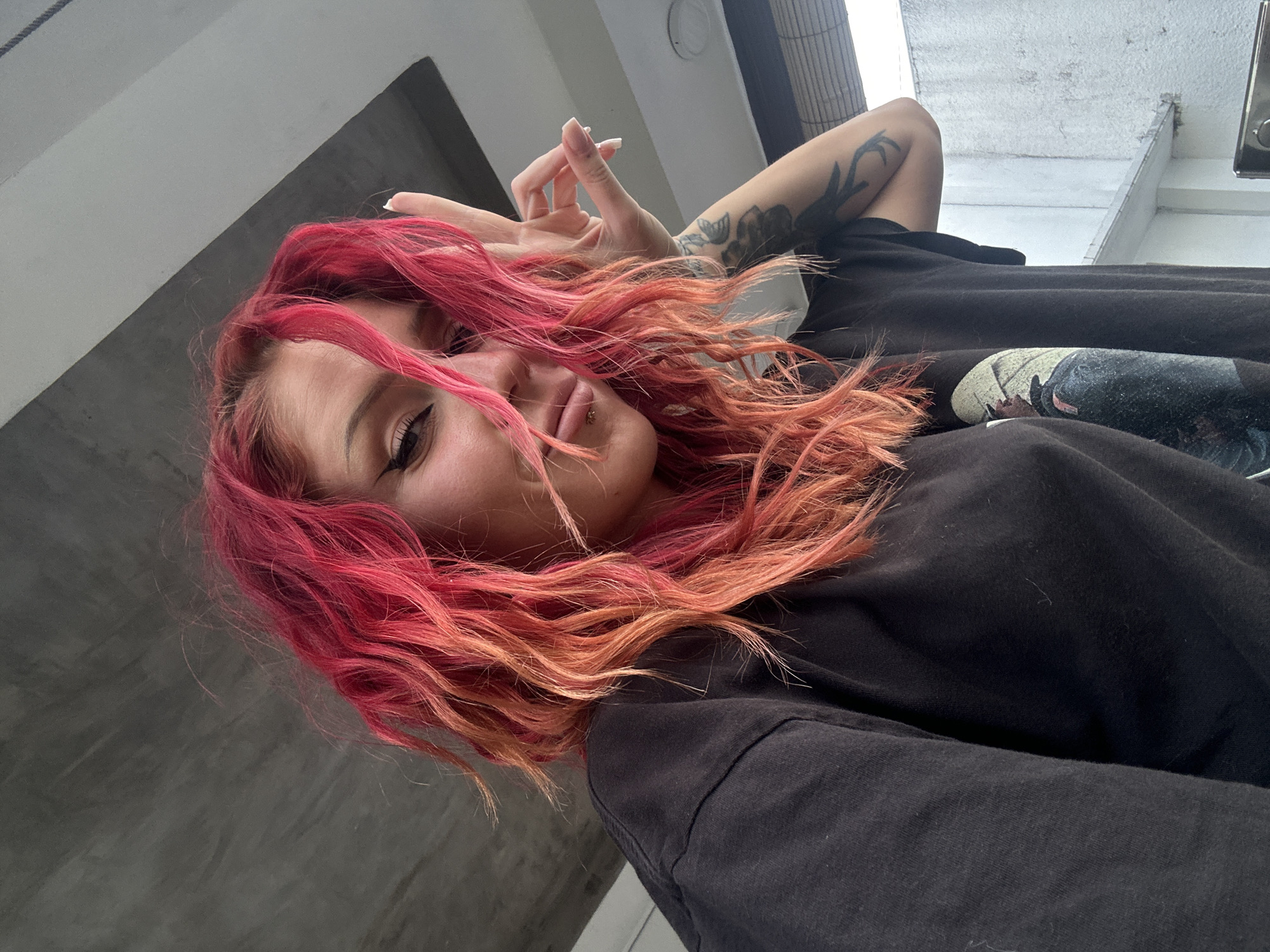 A low-angle, rotated indoor shot shows a young fair-skinned person with long, wavy red-to-orange ombre dyed hair. She is making a peace sign with her right hand, which features long manicured nails. She is wearing a black t-shirt. She has a lip piercing on the right side of her lower lip and apparent tattoos on her left arm. The background includes a gray wall on the left and a window with light streaming through from above on the right. A section of white ceiling is visible at the top right corner. The location, North Kuta, Indonesia, is not discernible from the provided image.