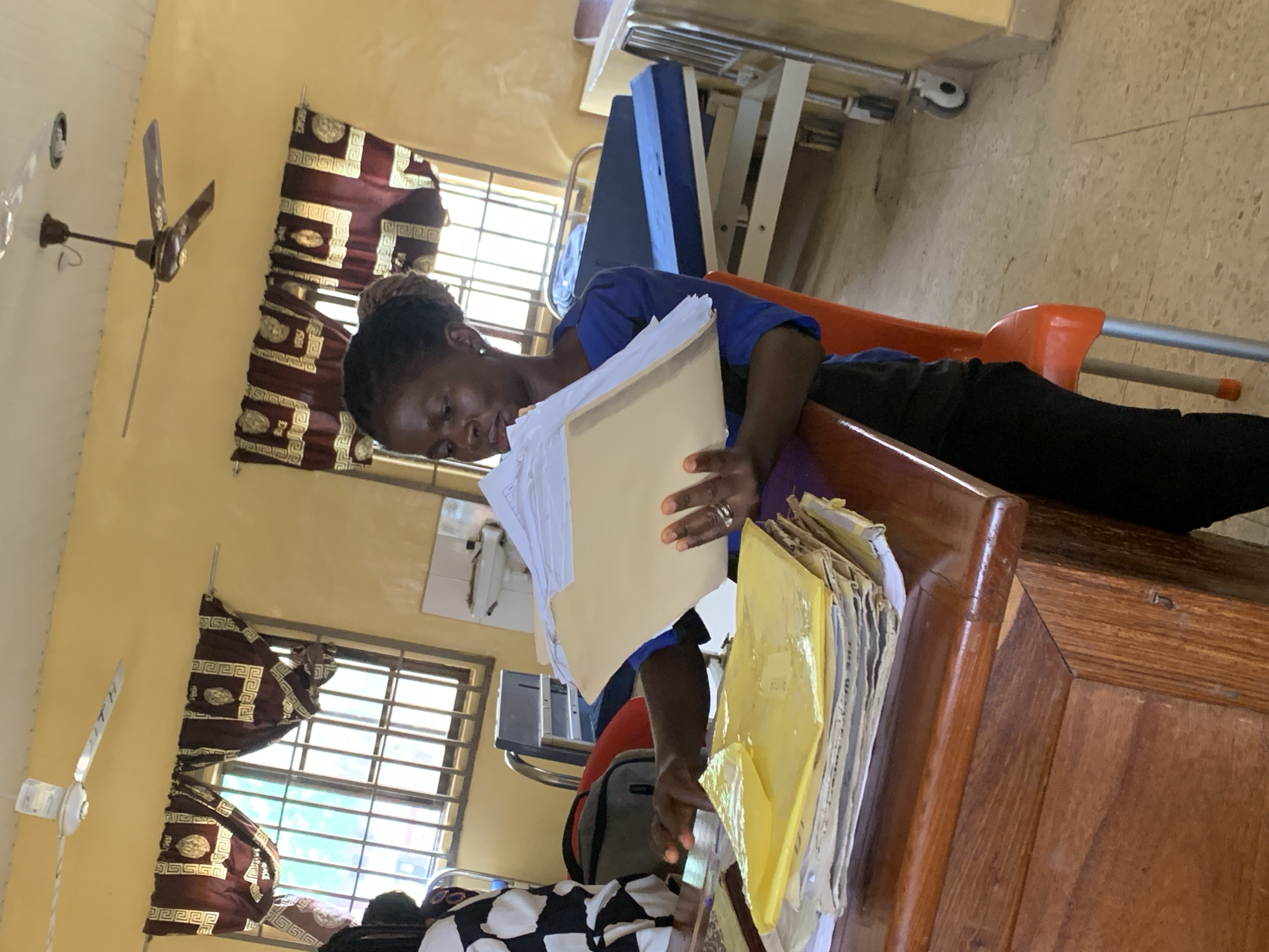 A woman is seated at a wooden desk in a room in Oko Erin, Nigeria. She is reviewing paperwork, holding a large file in her hands. The room features windows with burgundy patterned curtains, a ceiling fan, a bed, and a cart. The woman is wearing a blue shirt and black pants. Additional files are stacked on the desk.
