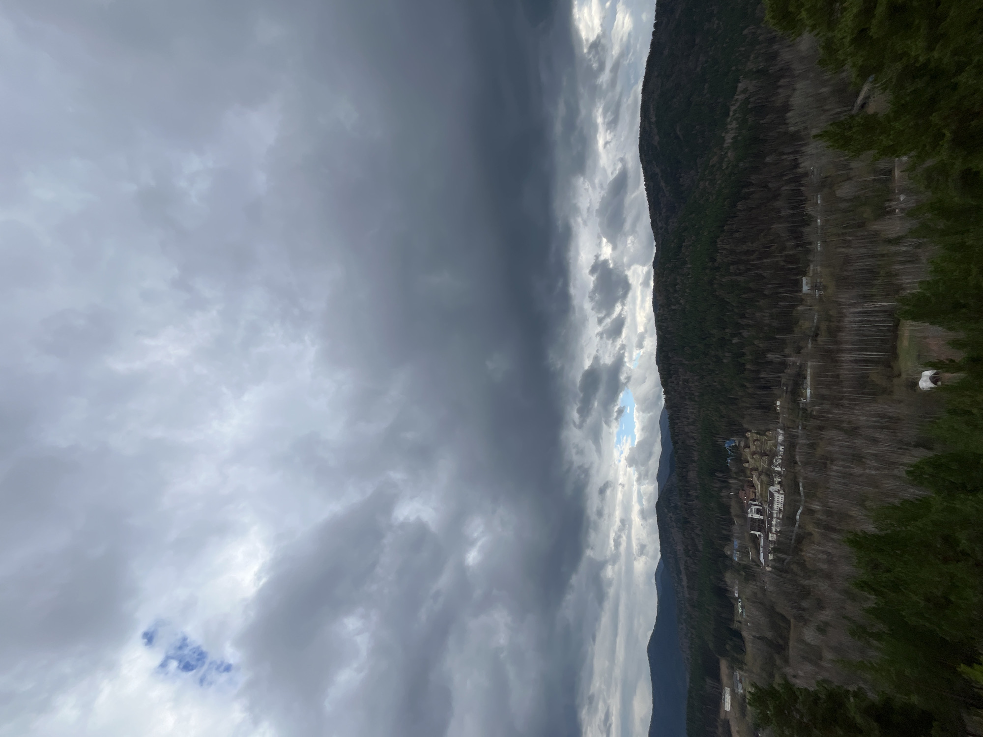 The image shows a view of Stolby Nature Reserve near Krasnoyarsk, Russia. The scene is dominated by a dramatic sky with heavy, dark storm clouds on the left side of the frame, contrasting with brighter, more scattered clouds on the right. A small patch of blue sky is visible between the clouds. Below the clouds, a forested mountainside is partially visible, with a small settlement or village nestled near the base. The vegetation on the mountain appears to be a mix of deciduous and coniferous trees, reflecting the transition to spring. The photo is taken from an elevated position, capturing the vastness of the natural landscape.