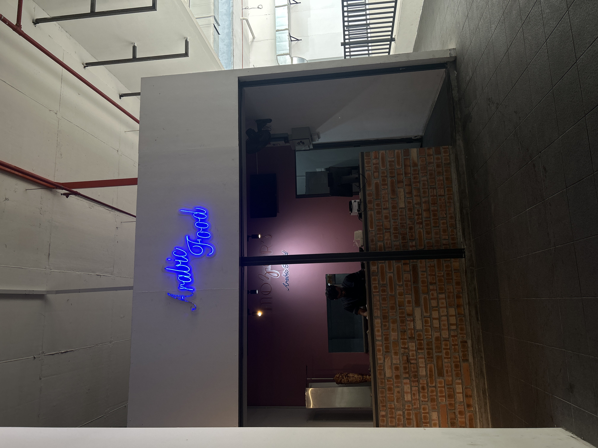 This image depicts the exterior of an Arabic food establishment, likely a restaurant or takeaway shop, located in an urban setting in Kuala Lumpur, Malaysia. The establishment features a modern facade with a combination of white and exposed brick walls, and a large glass storefront. Above the entrance, a neon sign in bright blue reads "Arabic Food" in a cursive font.
Inside the shop, the space is illuminated by warm lighting, highlighting the brick interior and the dark-colored walls. A person, appearing to be an employee, is visible behind the counter, wearing a dark uniform and a mask, suggesting the ongoing COVID-19 pandemic or general hygiene practices. They are engaged in what seems to be food preparation, with a vertical spit of meat, typical for shawarma, visible in the background.
The scene appears to be captured during the day, possibly in the late afternoon or early evening, given the lighting conditions and the shadow play. The setting is an alleyway or a walkway between buildings, with tiled walls and overhead structures suggesting an indoor or covered outdoor area. The overall impression is of a clean, contemporary food business catering to a local clientele. The composition is slightly tilted, adding a dynamic perspective to the scene.