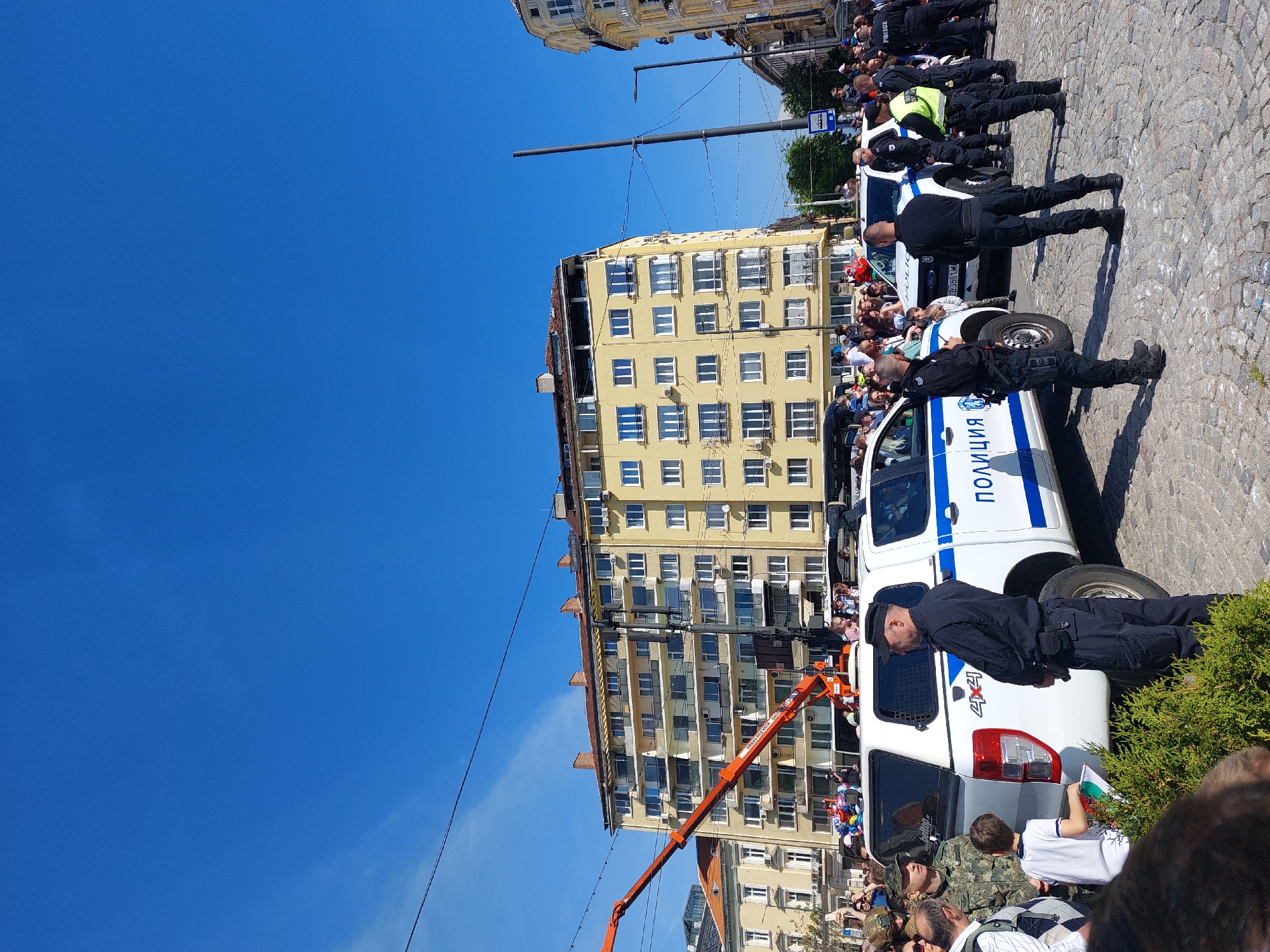 The image depicts a crowd of people gathered on a cobblestone street in what seems to be a city, possibly in Bulgaria based on the presence of vehicles marked with "Полиция" (Police in Bulgarian). There is a tall, yellow building on the left, with rows of windows. An orange crane is extended in front of the building. Police officers are standing near white police vehicles. The sky is clear and blue.