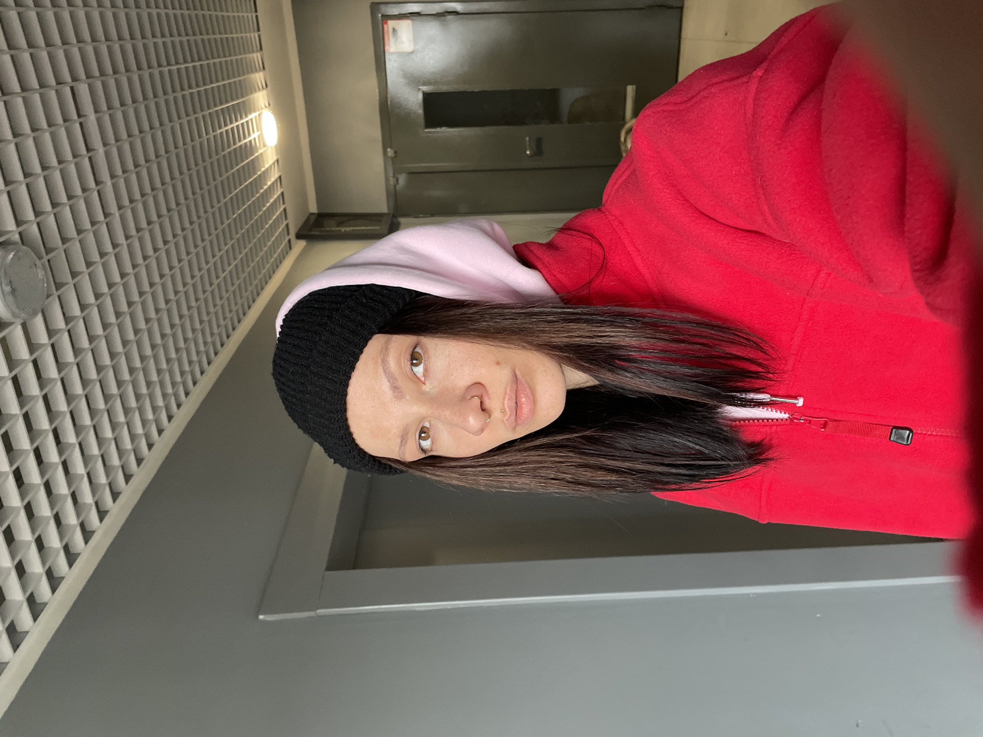 A woman in a black knit hat, pink hoodie, and red zip-up jacket is indoors. The location appears to be a hallway with a tiled ceiling and a dark metal utility box. The image lacks features clearly identifiable as Izhevsk, Russia.
