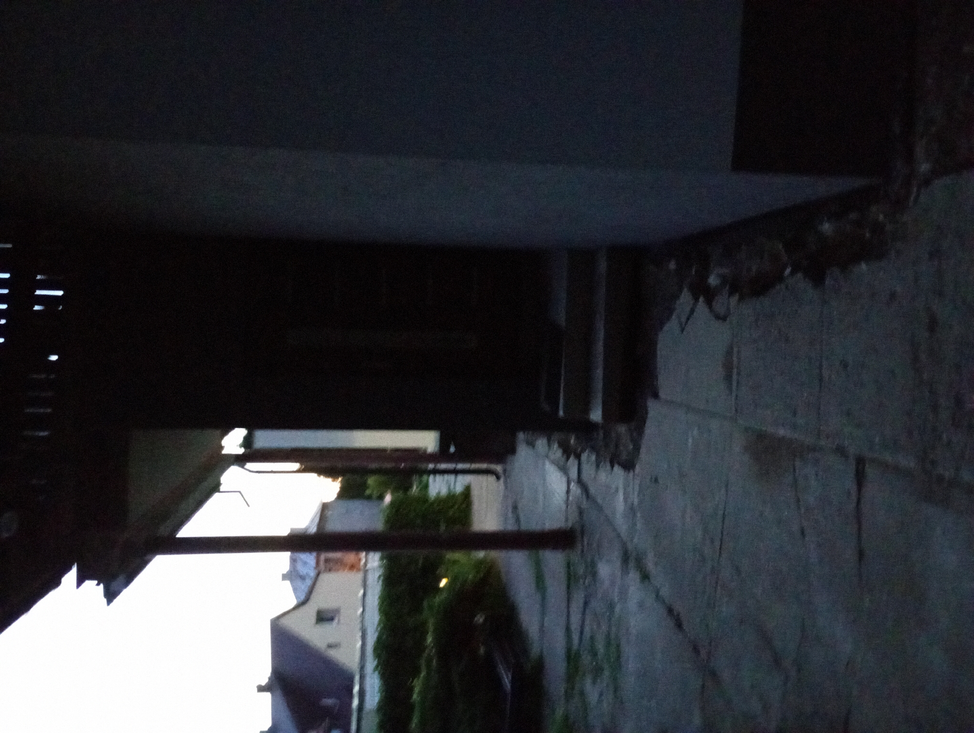 The image depicts a dimly lit outdoor scene, possibly in the city of Białystok, Poland, at dusk. A concrete pathway, marked with cracks and appearing slightly wet, runs alongside a dark building and a dense hedge. A covered walkway with supporting posts extends from the building into the frame. In the background, residential buildings with pitched roofs are visible under a pale sky. The scene suggests an exterior residential or commercial area, emphasizing shadows and low light conditions.