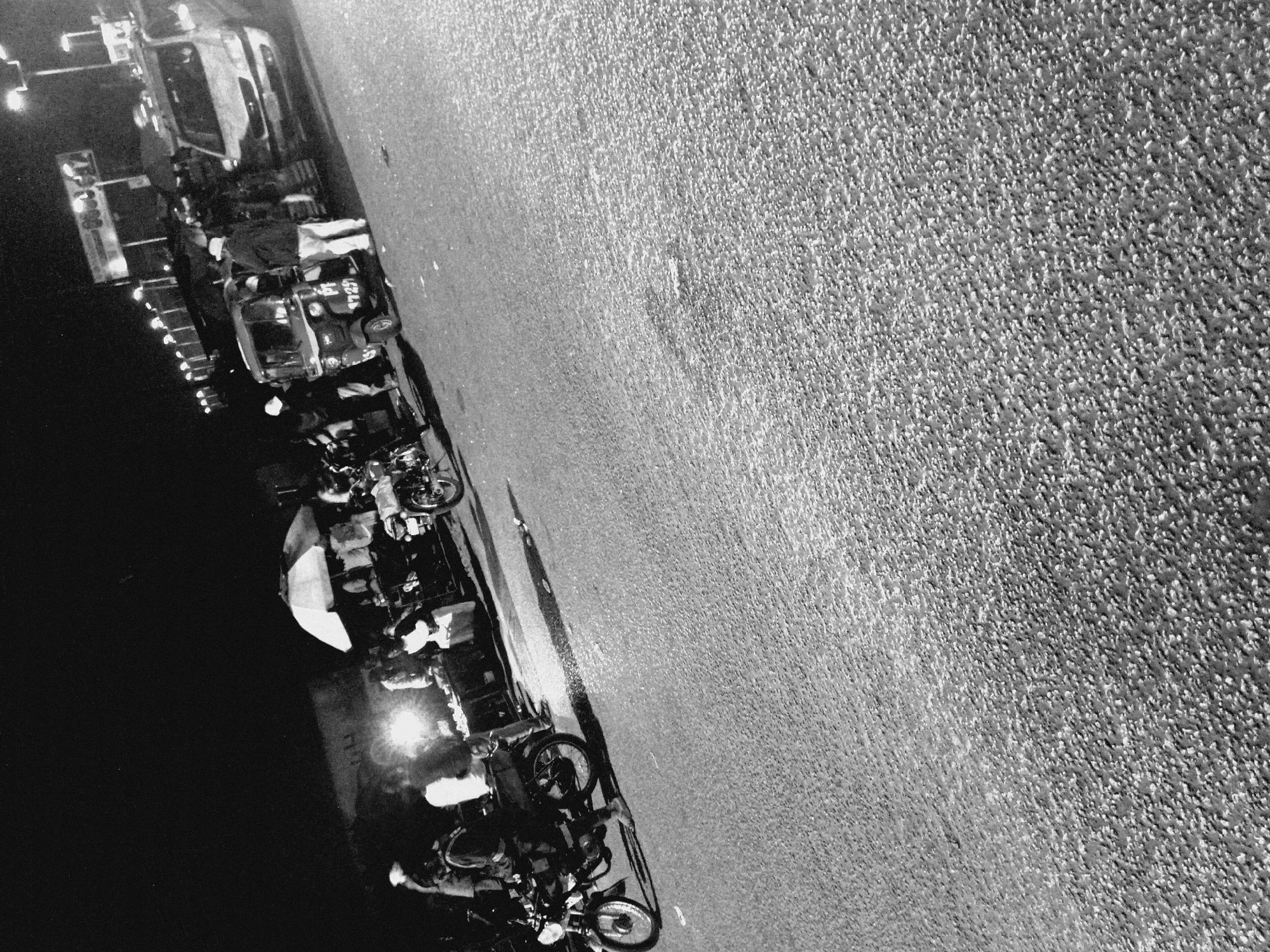 The image depicts a nighttime street scene in Bukuru, Nigeria, with a focus on a busy roadside market. In the foreground, a motorcycle rider is positioned next to a stall. Several other motorcycles are visible further back. A tuk-tuk, also known as a "Keke Napep," is parked near a food stall, which is illuminated by a bright light and covered by a large umbrella. A white van is also present in the background, suggesting vehicular traffic. The market appears to be active, with several people gathered around the stalls, likely engaged in commerce or social interaction. The overall environment suggests a lively urban setting at night, characterized by street vendors and transportation.