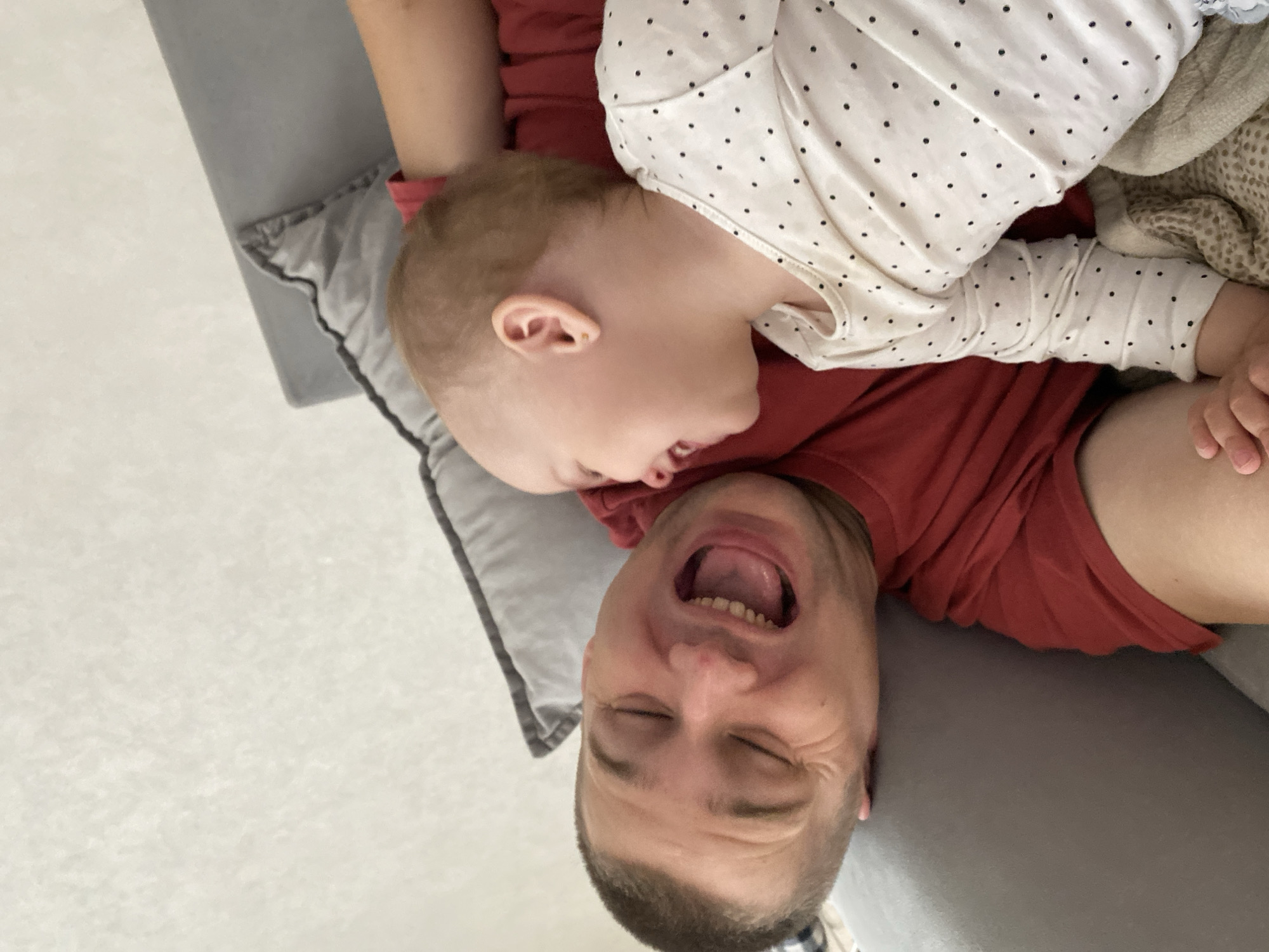 An indoor photograph depicts a light-skinned adult male and a light-skinned infant/toddler engaged in playful interaction, situated on a grey sofa in what is presumably a residence in Chaikovsky, Russia. The male, appearing to be in his 30s-40s with short brown hair, lies on his back, wearing a red short-sleeved t-shirt. His head is tilted towards the right, eyes are closed, and his mouth is wide open in an expression of laughter or joyous exclamation. Visible wrinkles are present around his closed eyes. The infant/toddler, positioned to the male's right and facing him, has very short light brown/blonde hair and wears a white long-sleeved top with small black polka dots. A small gold stud earring is visible in the child's right ear. The child's mouth is open in a smile, and eyes are closed or nearly closed, reflecting a similar state of joy. The child's left arm rests on the male's chest/torso. A grey throw pillow with dark piping is visible behind the male's head. A textured light-colored blanket and a patterned blanket are partially visible beneath the child. The background consists of a plain light-colored wall.