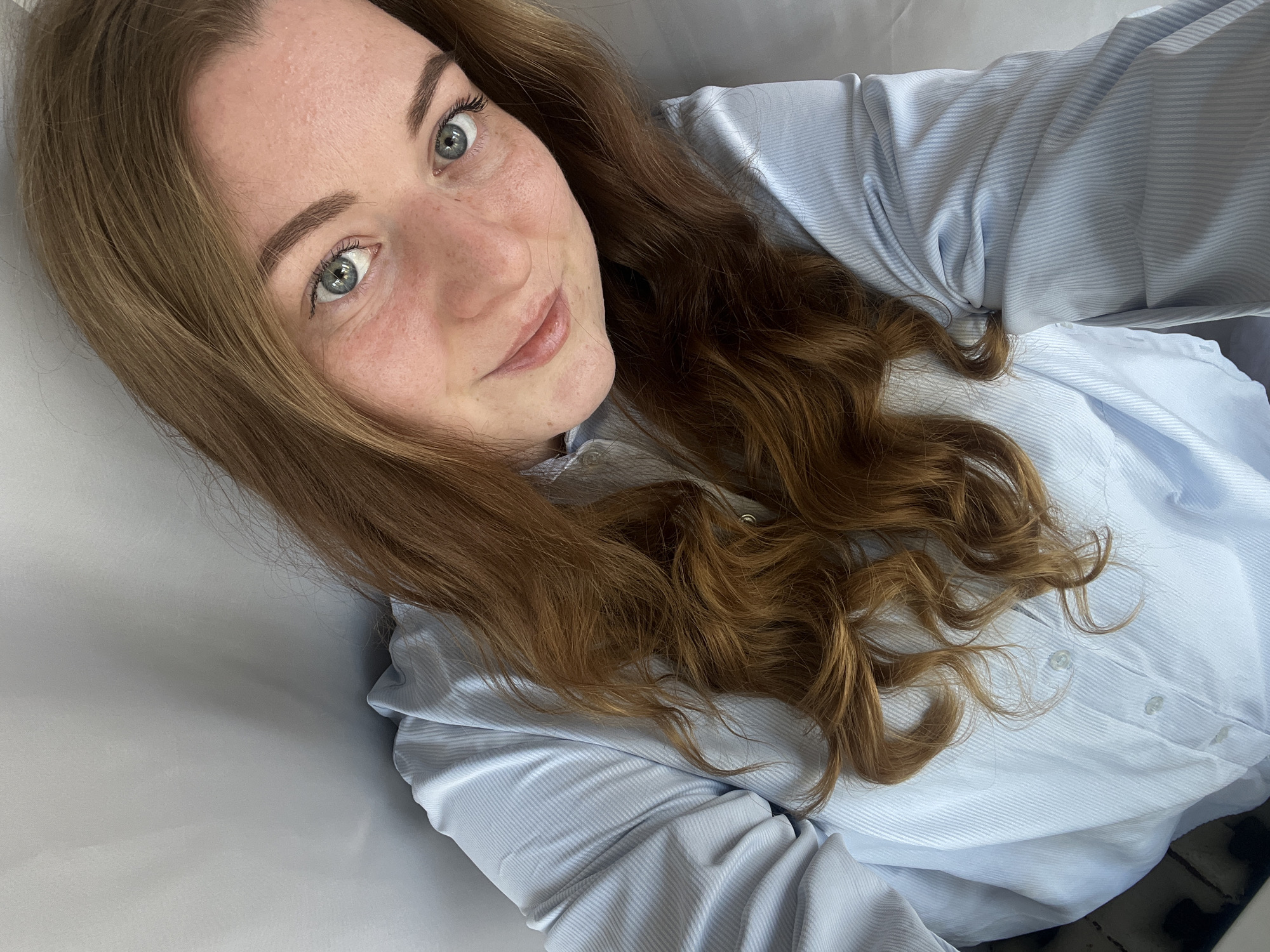 A young woman with auburn hair and light eyes is lying on a white surface, possibly a bed or a couch. She is wearing a light blue, button-down shirt. The camera angle is from above, suggesting a selfie. Her expression is relaxed and slightly smiling. The location of Раздолье, Russia, is not evident within the image itself.