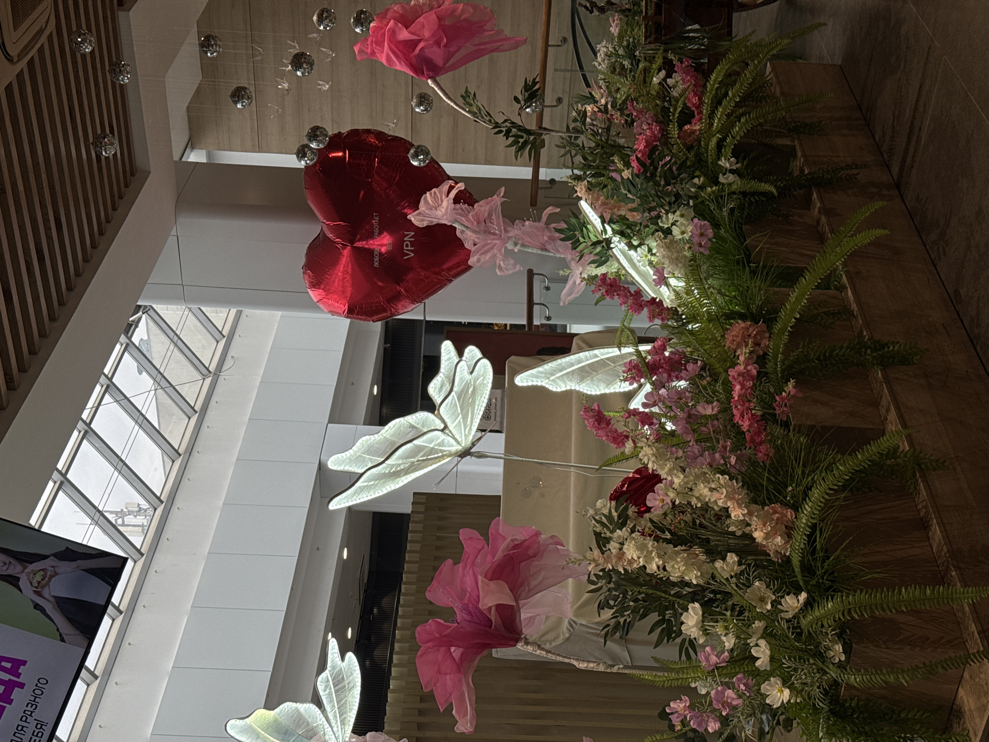 The image displays an ornate indoor decorative display within a modern public building, likely a shopping mall in Izhevsk, Russia. The perspective is upward, showcasing an elaborate arrangement of artificial flowers, illuminated elements, and balloons hanging from a high ceiling.
The central feature is a vibrant, artificial floral display, rich with pink, white, and red blossoms, complemented by lush green foliage including ferns. Several large, translucent white butterfly cutouts, which appear illuminated, are integrated into the arrangement, creating a whimsical atmosphere. Two glossy red heart-shaped foil balloons are prominent, with one bearing Cyrillic text: "ЛЮБОВЬ РАБОТАЕТ" (LOVE WORKS) and "VPN" below it. Delicate pink, sheer fabric bows and large, soft pink, paper-like flowers further enhance the suspended décor.
Architectural elements include a high, white ceiling from which numerous small, spherical, reflective ornaments hang. Large skylight windows with geometric frames are visible, allowing natural light to illuminate the space. A wooden slatted wall or railing is seen on one side. In the lower left, a large digital display screen shows a partial image of a person holding food, with fragmented Cyrillic text including "НА" and parts of "...ДЛЯ РАЗНОГО ...ЕБЯ!".
The decorations suggest a festive or celebratory occasion, possibly related to Valentine's Day or a general spring theme, taking place during the daytime. No people are directly visible in the scene.