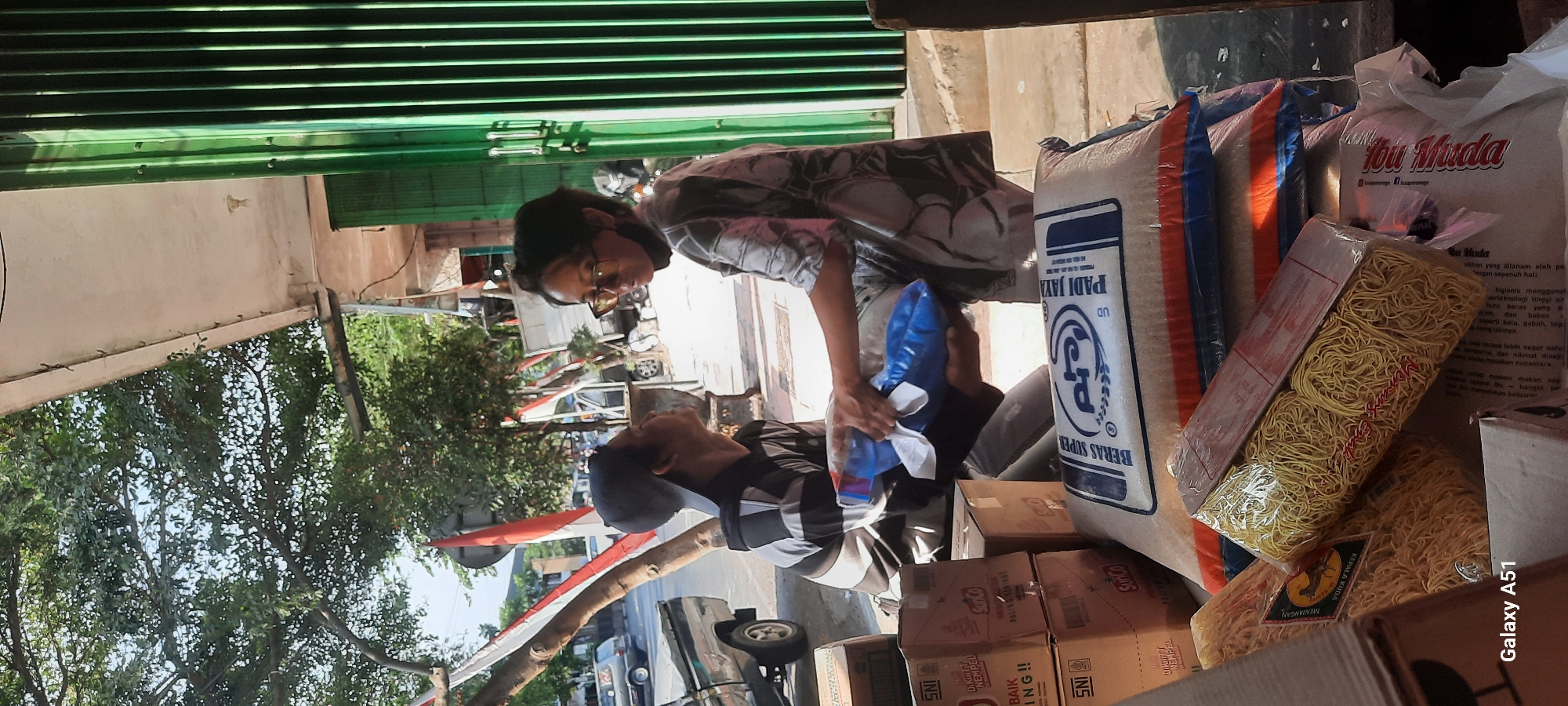 In Beru, Indonesia, two individuals are observed interacting on a street in front of a building. One person, wearing a cap and a striped shirt, is seated and looking upwards. The other person, wearing glasses and a patterned shirt, is standing and handing a blue bag to the seated individual. In the foreground, various goods are displayed, including boxes, bags of rice, and packages of instant noodles. The background shows a street scene with vehicles and trees, suggesting an outdoor commercial area. The overall impression is a transactional moment, possibly a delivery or exchange of goods.