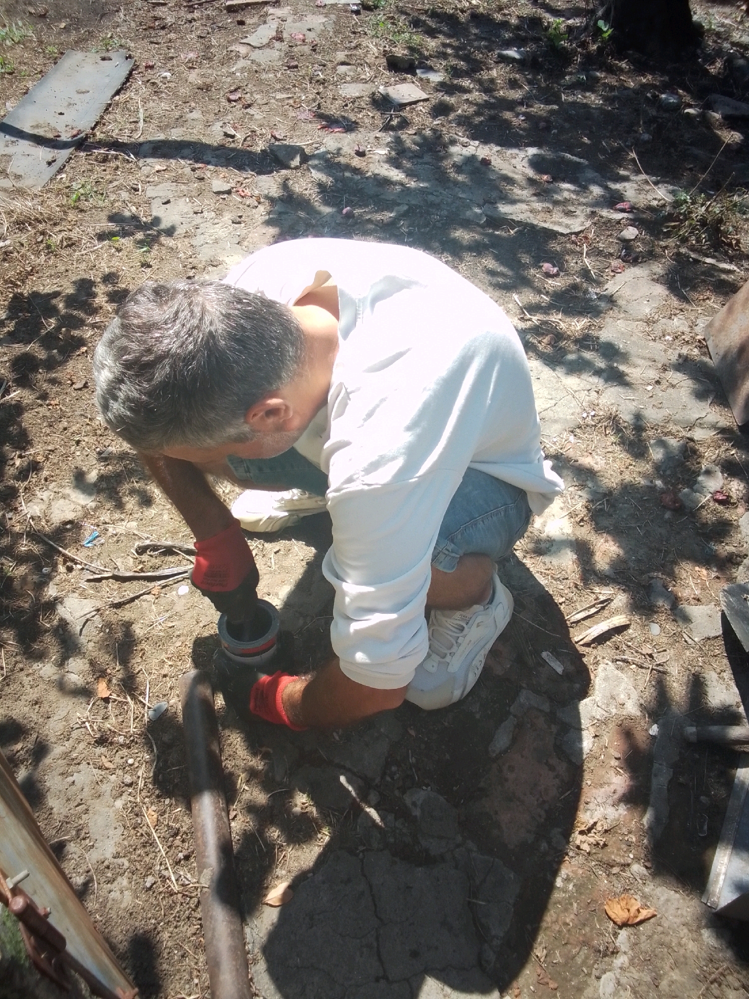 A man with grey hair is kneeling on the ground, wearing a white long-sleeved shirt, blue denim shorts, red gloves, and white sneakers. He is holding a grey pipe fitting with a red ring in his gloved hands and appears to be working on it. The ground around him is a mixture of dirt, small rocks, and dry grass. There are shadows cast from overhead foliage, suggesting an outdoor environment, possibly a yard or a construction area. The background shows more of the dry, dusty ground with some scattered debris. The scene does not provide any specific visual cues to pinpoint its location in Tbilisi, Georgia.