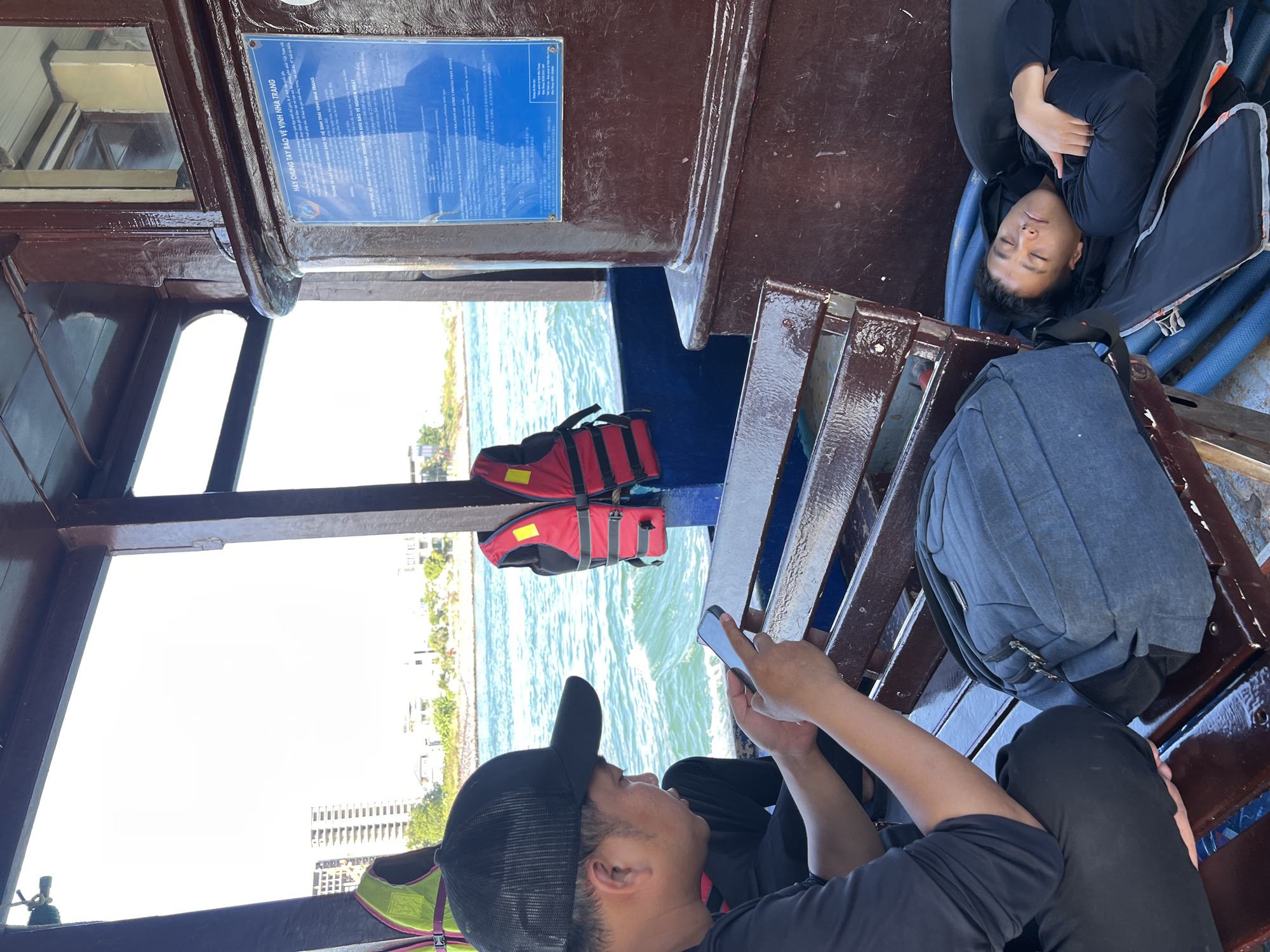 The image shows the interior of a boat on the water, with the coastline of Nam Nha Trang Ward, Vietnam, visible through the boat's windows. One man in a black shirt and cap is looking at and interacting with a smartphone. Another man is asleep in a sleeping bag on a bench in the boat. Life vests are placed on the boat's railing, and a backpack sits on the bench next to the seated man. A blue information plaque is mounted on the wall. The scene suggests a leisure or travel setting on the water.
